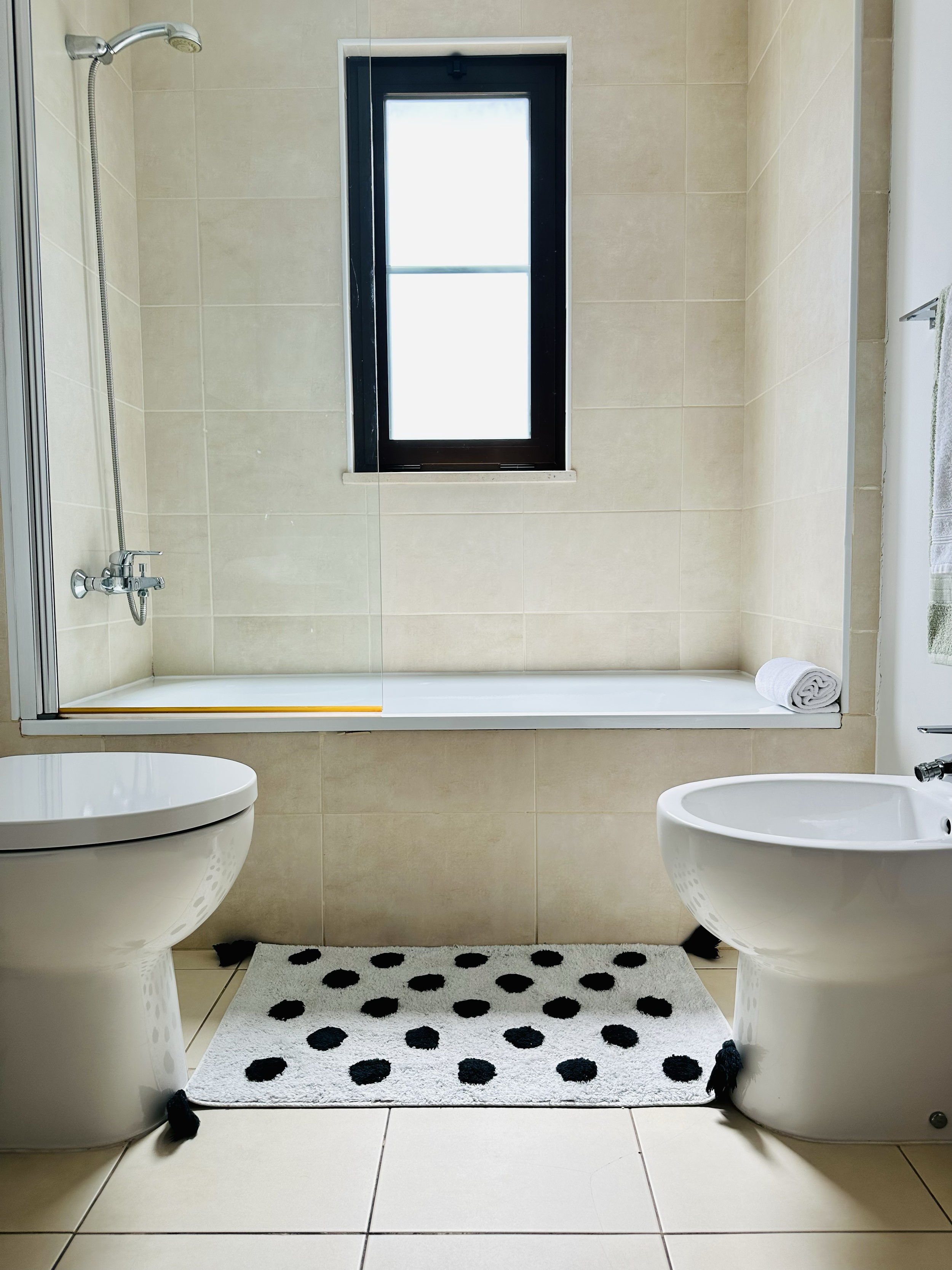 Bathroom with a window, showerhead, toilet, bidet, and a white rug with black polka dots.