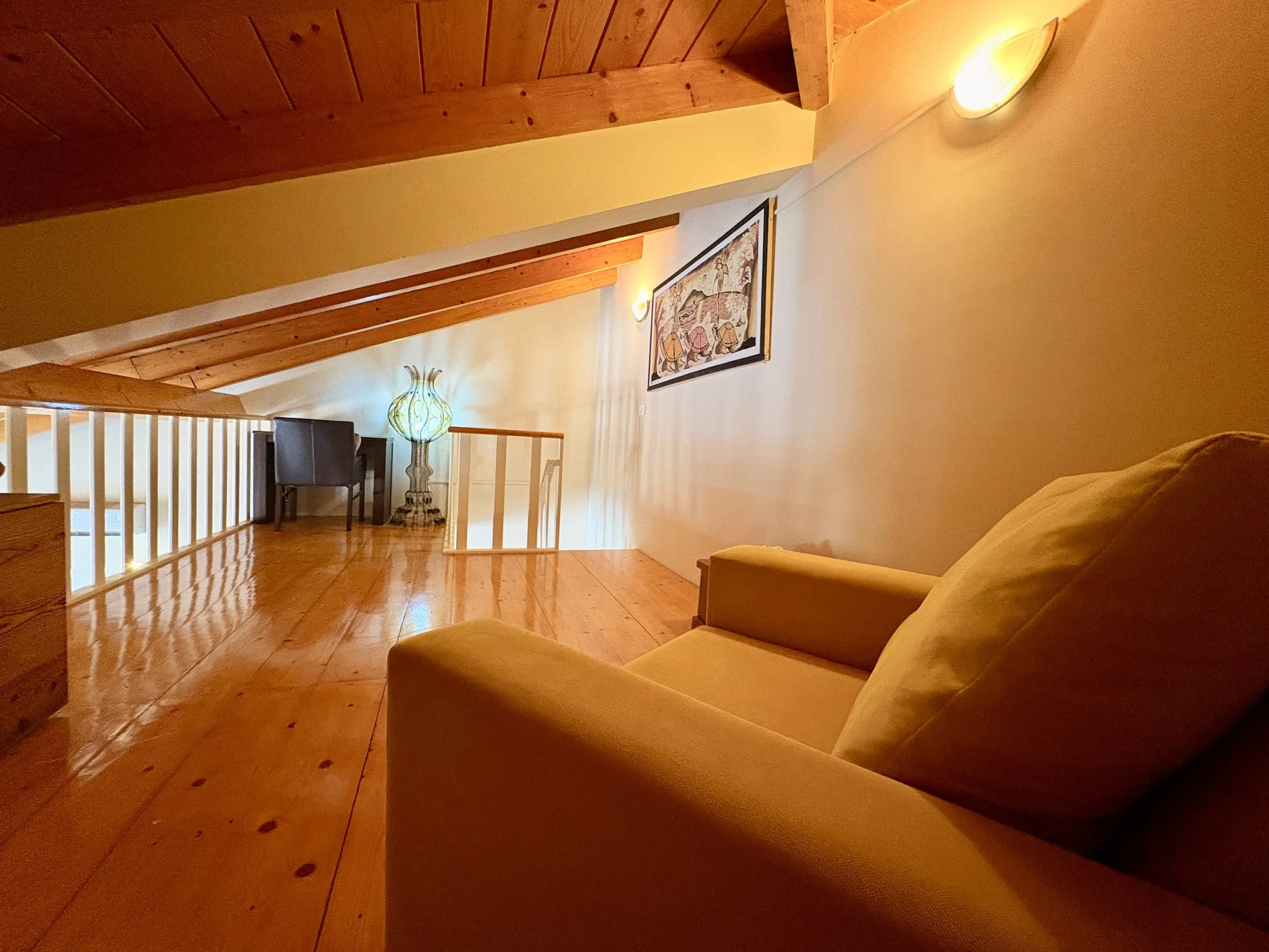 A cozy living space with wooden beams on the ceiling, a framed artwork on the wall, a yellow couch, and warm lighting from wall sconces and a decorative lamp. The floor is hardwood, and there is a white railing to the lower level visible in the foreg