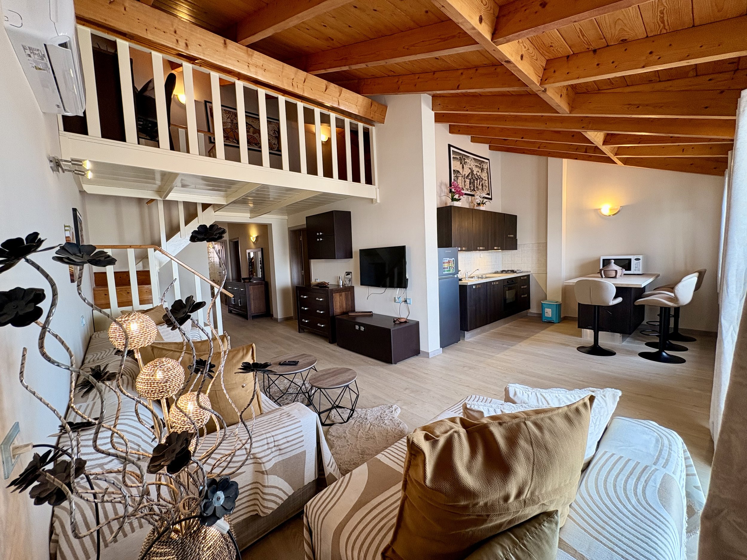 Living room with wooden vaulted ceiling, white walls, a loft railing, a tan sofa with pillows, a black TV stand with a flat-screen TV, a kitchen area with dark cabinets, a microwave, and a small dining area with stools. Decorative plants and artwork 