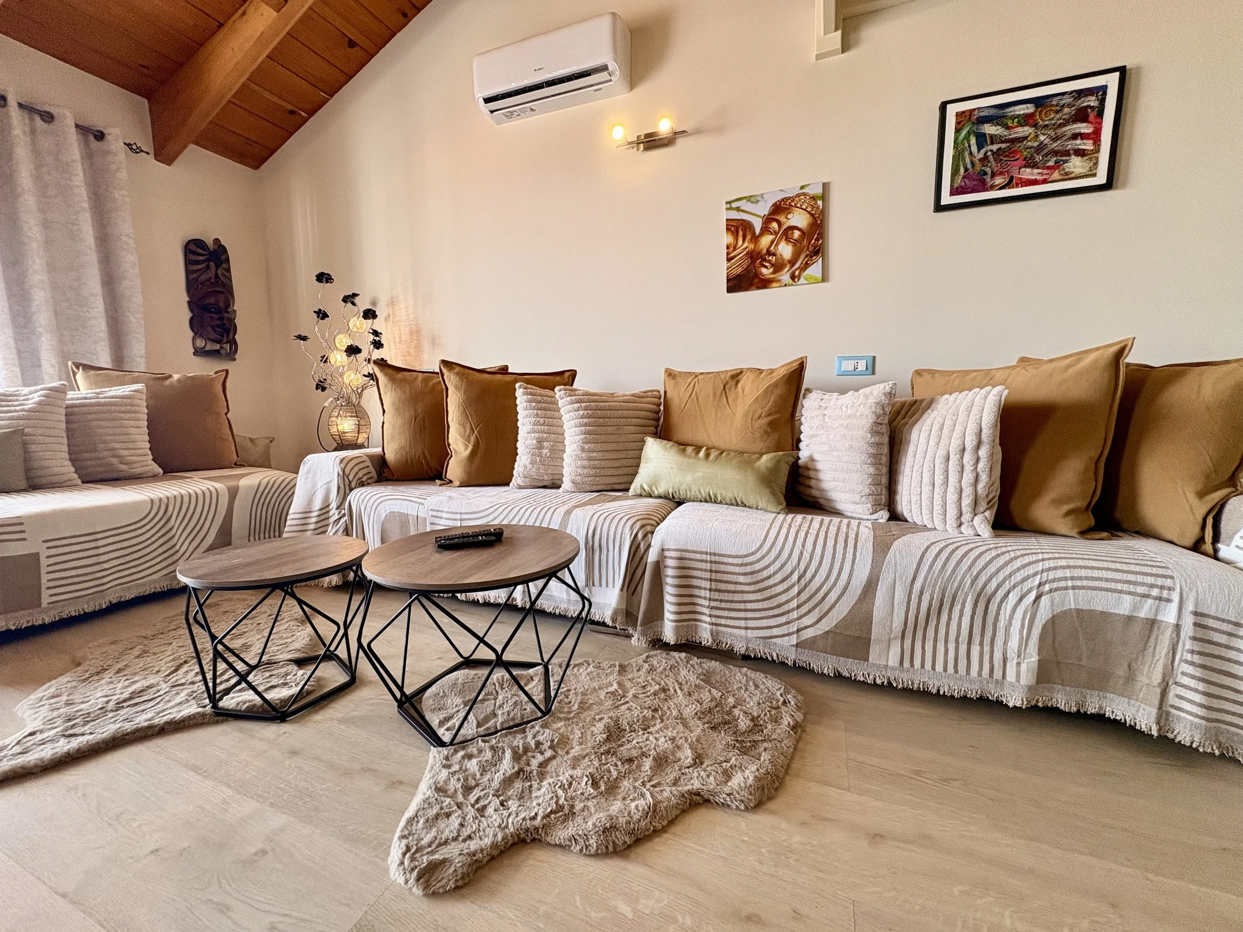 A living room with a beige sectional sofa decorated with various pillows, a round coffee table, a textured beige rug, wall art, a floor lamp, window curtains, and an air conditioning unit on the wall.