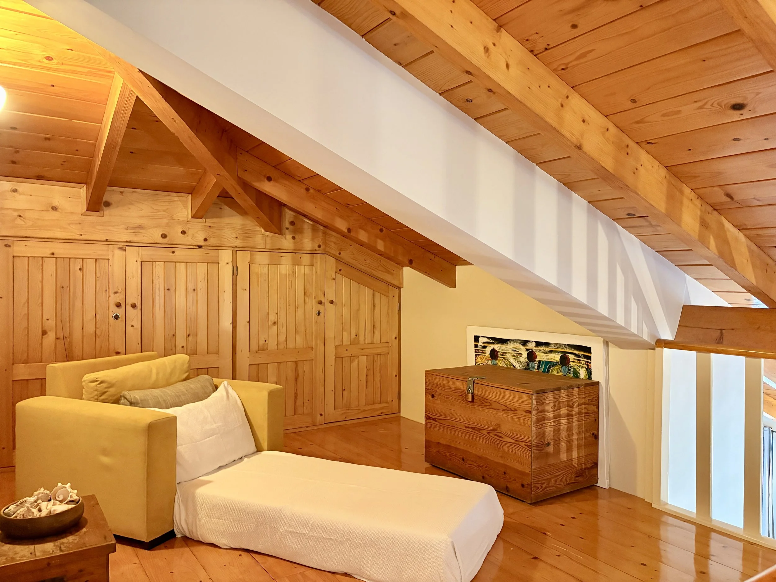 A cozy bedroom with wooden walls, ceiling, cabinets, and furniture, featuring a yellow armchair and a small wooden side table with seashell decor. There is a painting hanging on the wall and a staircase railing in the corner.