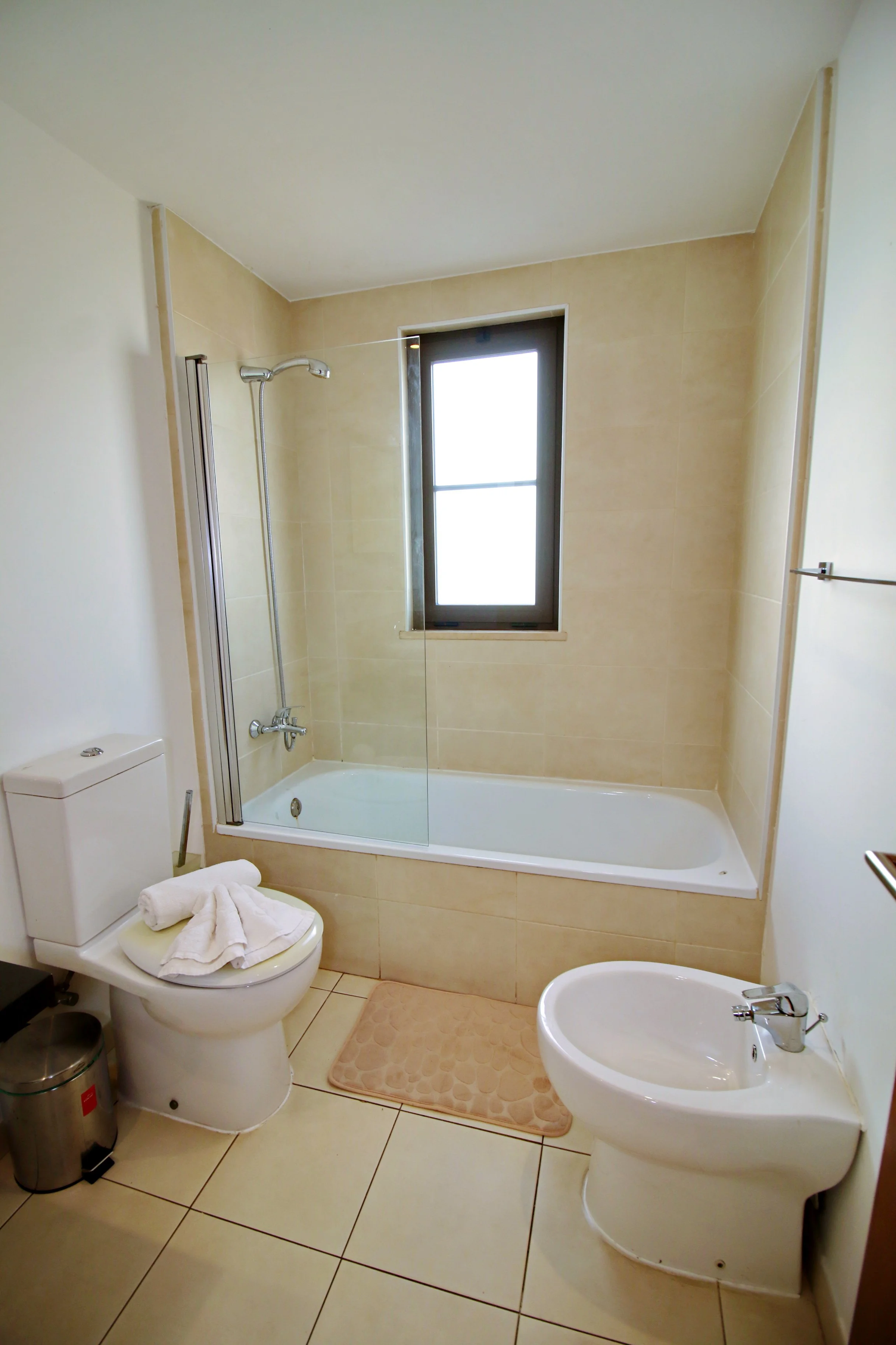 A bathroom with a bathtub, a showerhead, a toilet with towels, a bidet, and a window with frosted glass.