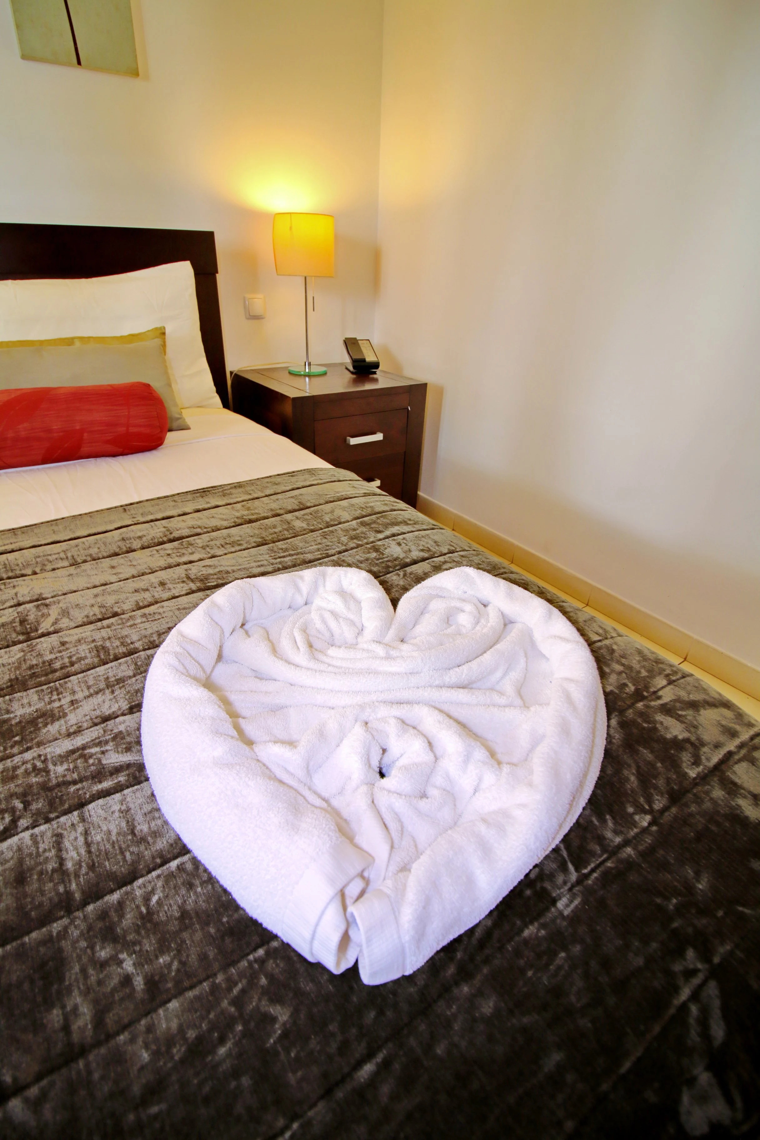 A hotel room with a bed, a nightstand, a lamp, and folded towels arranged in a rose shape on the bed.