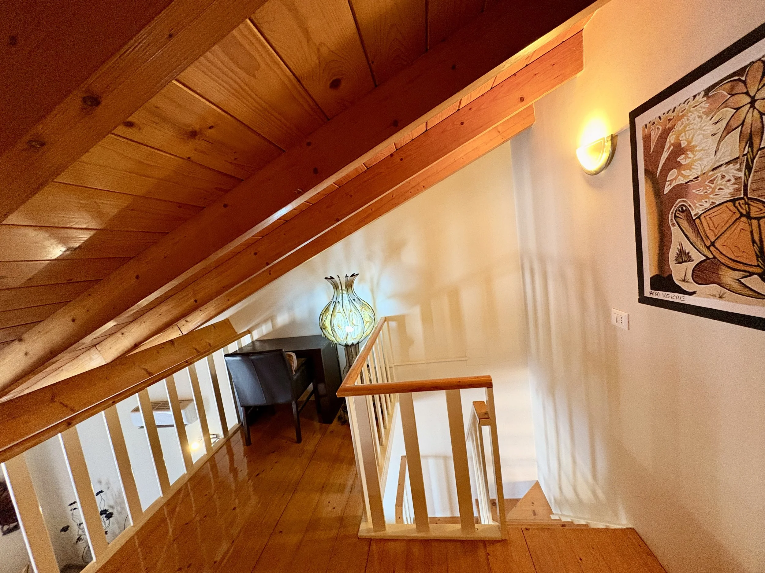 Interior view of a room with wood-paneled ceiling and staircase railing, illuminated by a wall sconce and a decorative glass lamp, with a framed artwork on the wall featuring a stylized image of a turtle and plant motifs.
