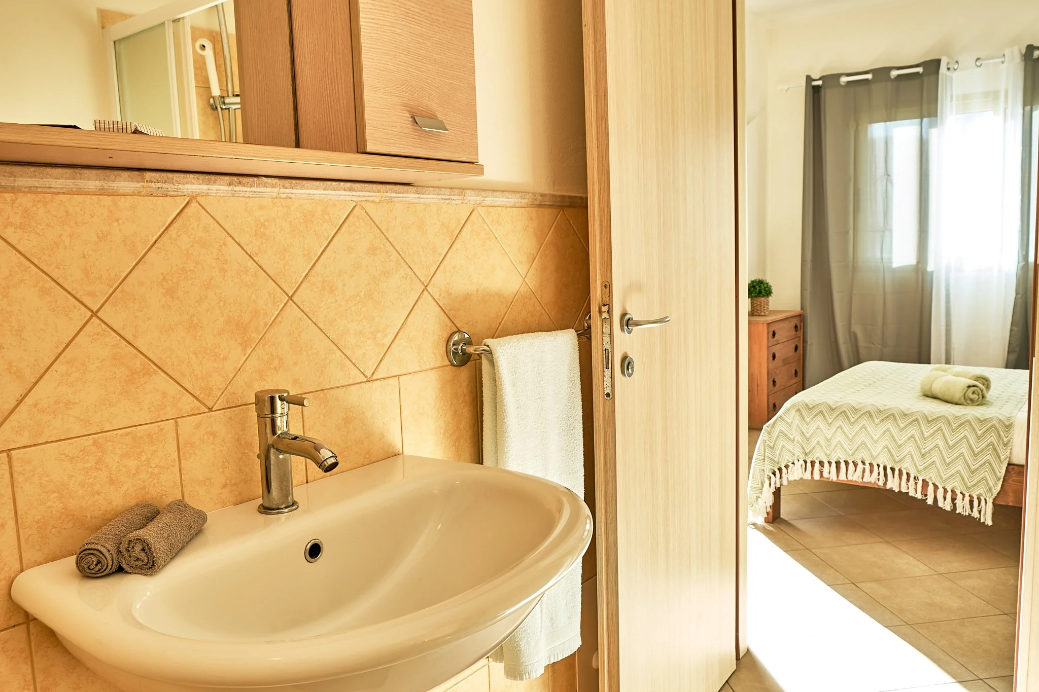 A bathroom with a sink, towel, and mirror, and a bedroom visible through a doorway with a bed, window with curtains, and a small dresser with a potted plant.