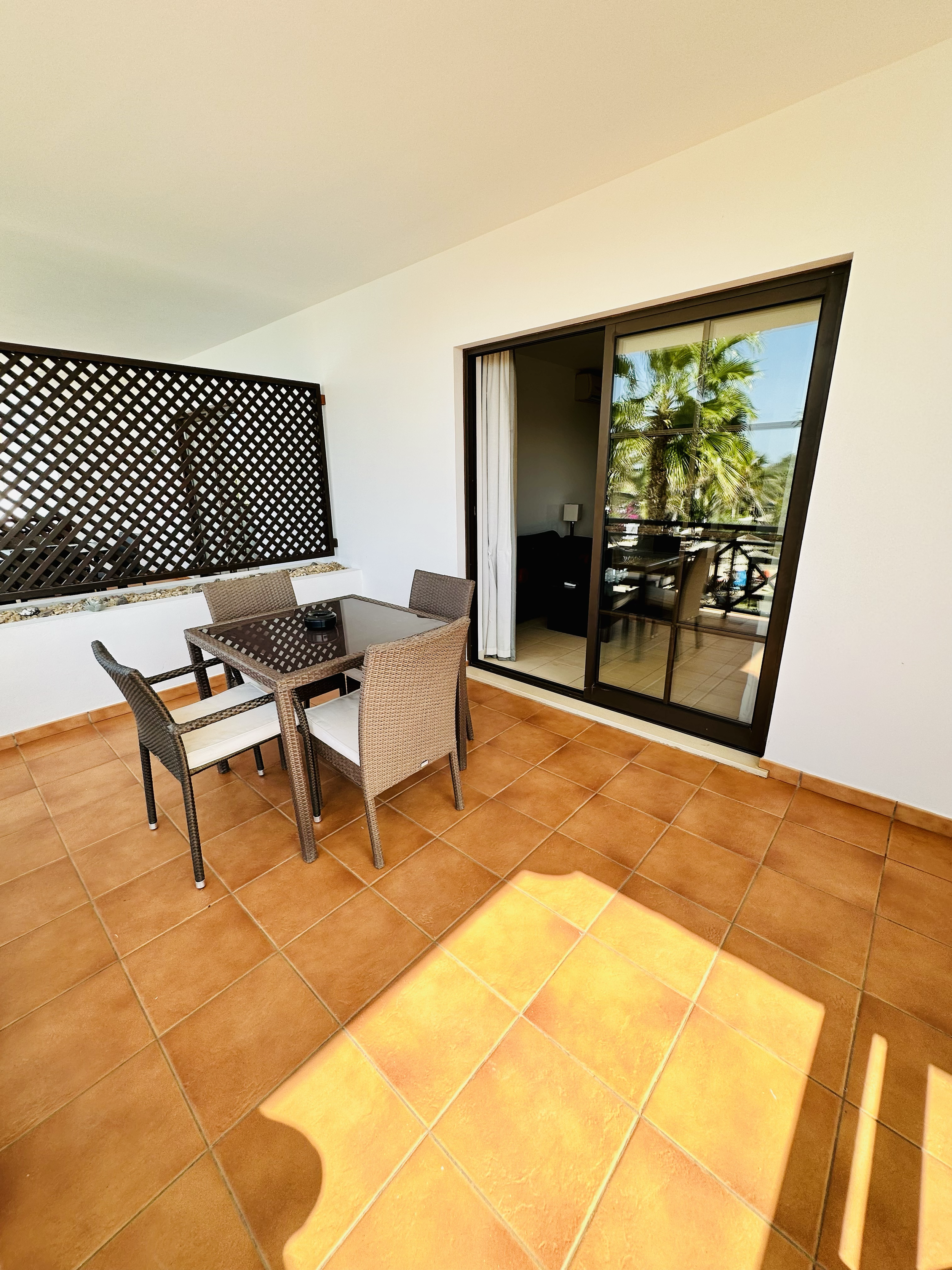 A balcony or patio area with brown tiled flooring, wicker chairs around a glass table, a wooden lattice privacy screen, and a sliding glass door with a view of palm trees outside.