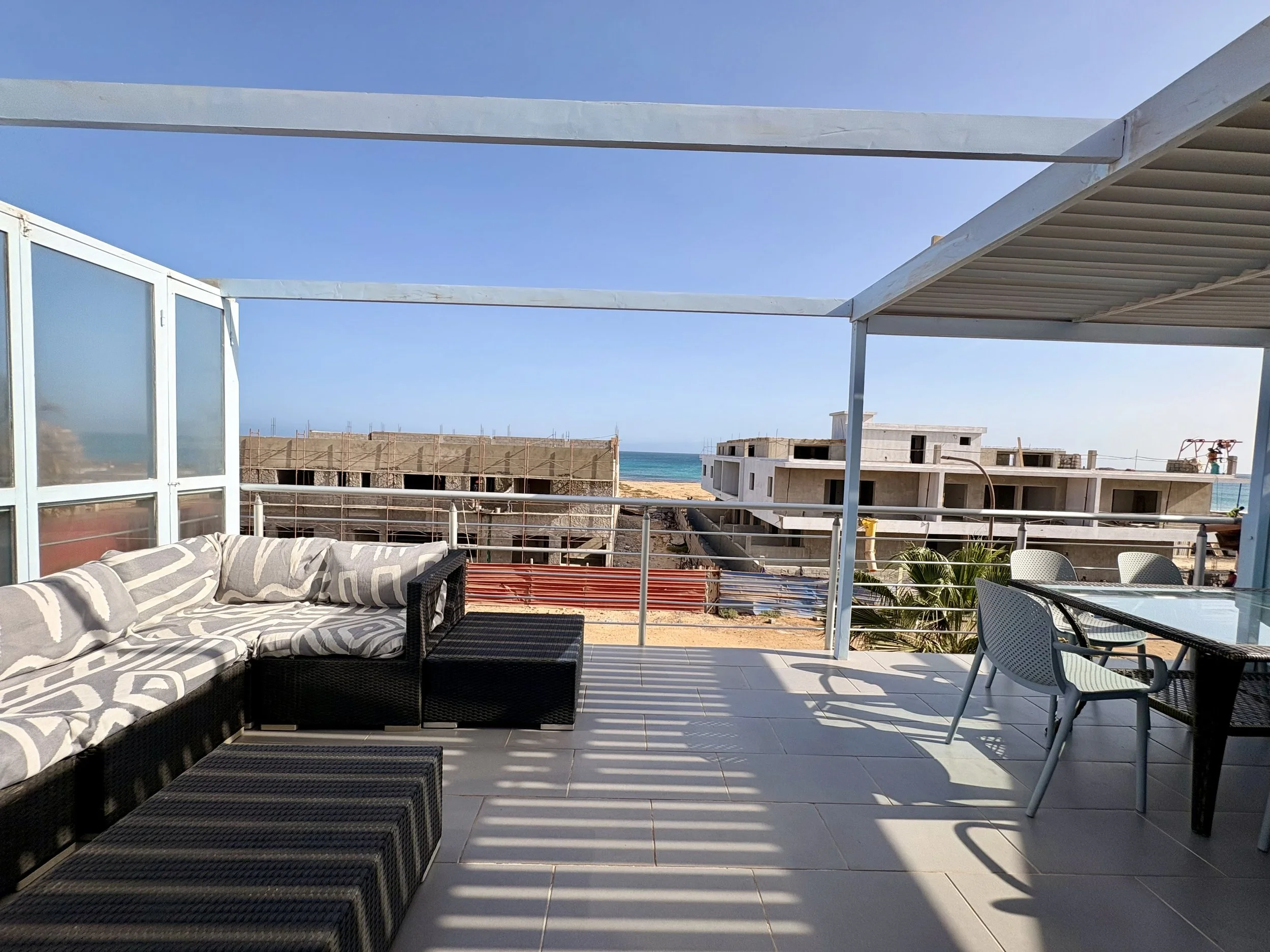 A balcony with outdoor furniture including a patterned cushioned sofa, black wicker table, and plastic chairs, overlooking a beach and the ocean under a clear blue sky.