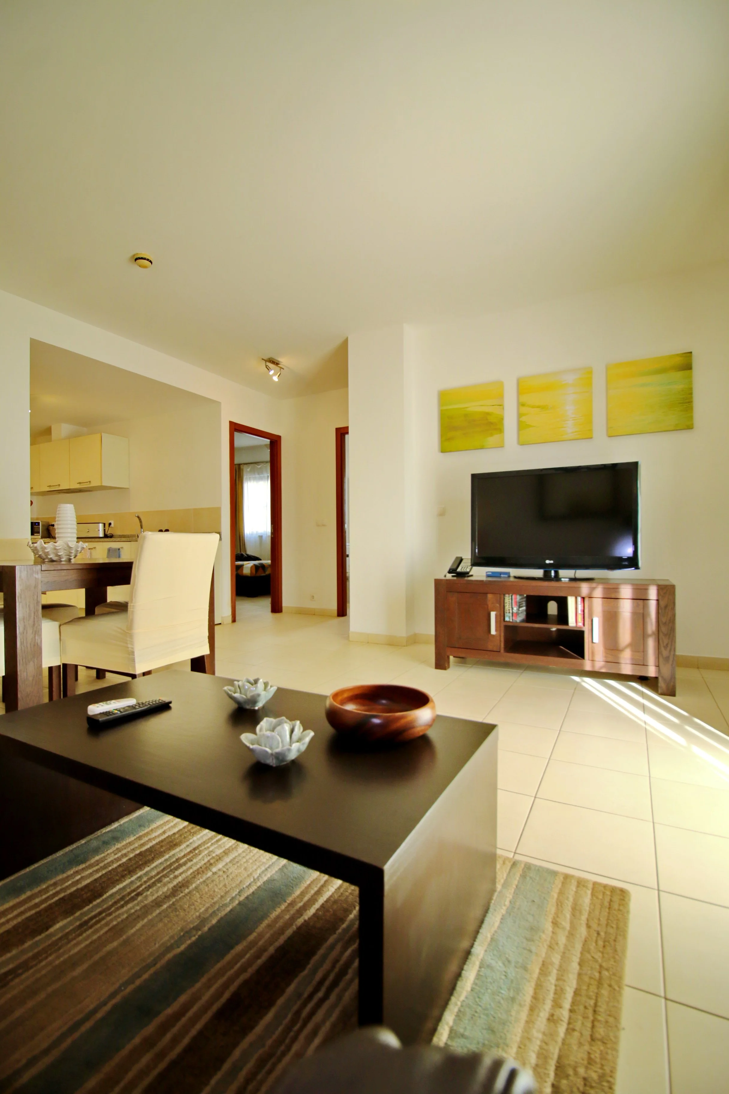Living room with a TV on a wooden stand, paintings on the wall, a dark coffee table with decorative bowls, a striped rug, and a dining area with chairs and a kitchen in the background.