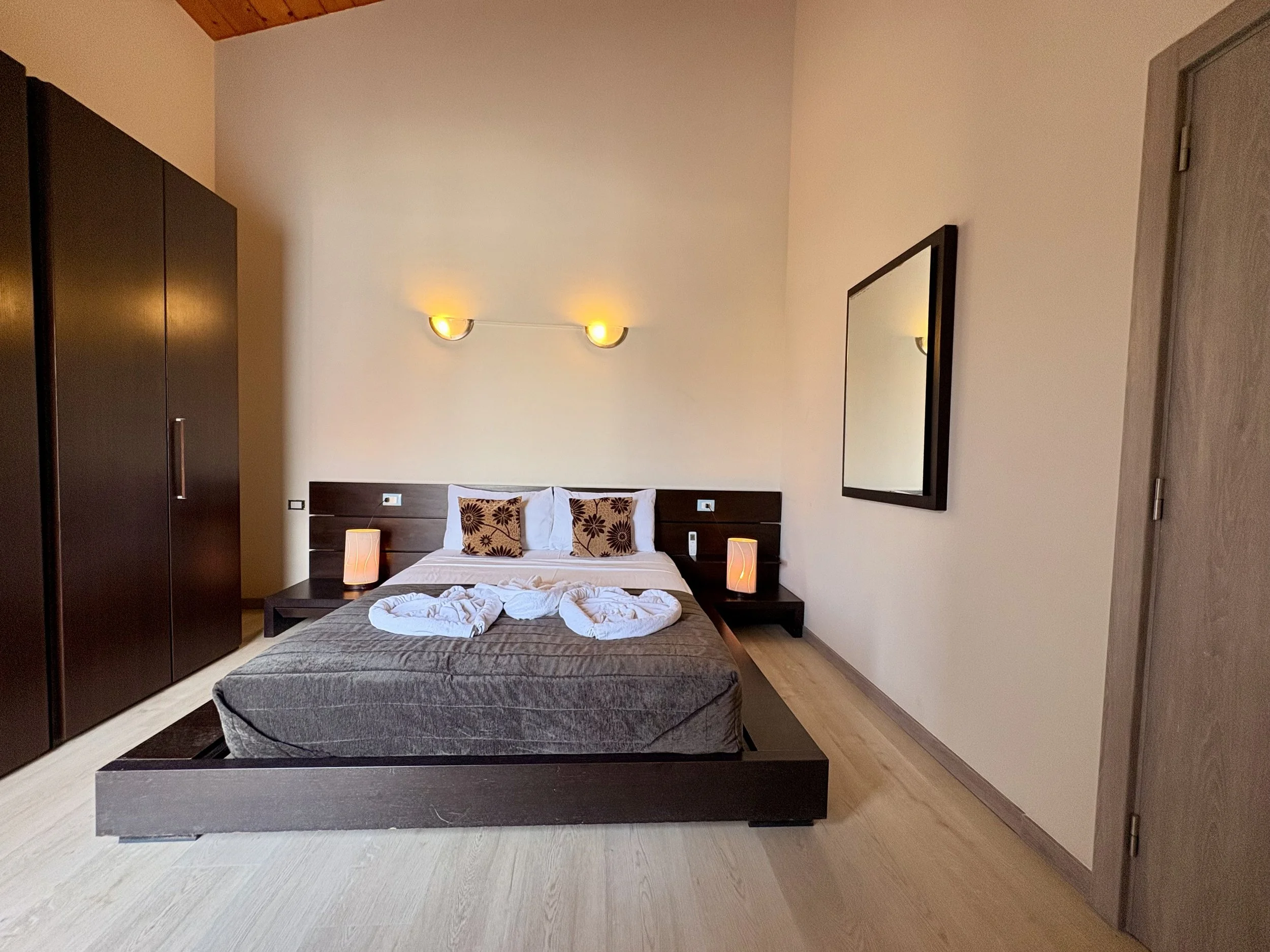 A bedroom with a bed in the center, a dark wooden headboard with two pillows, bedside tables with lamps, a large mirror on the wall, and a dark wardrobe on the left side. The room has light-colored flooring and beige walls.