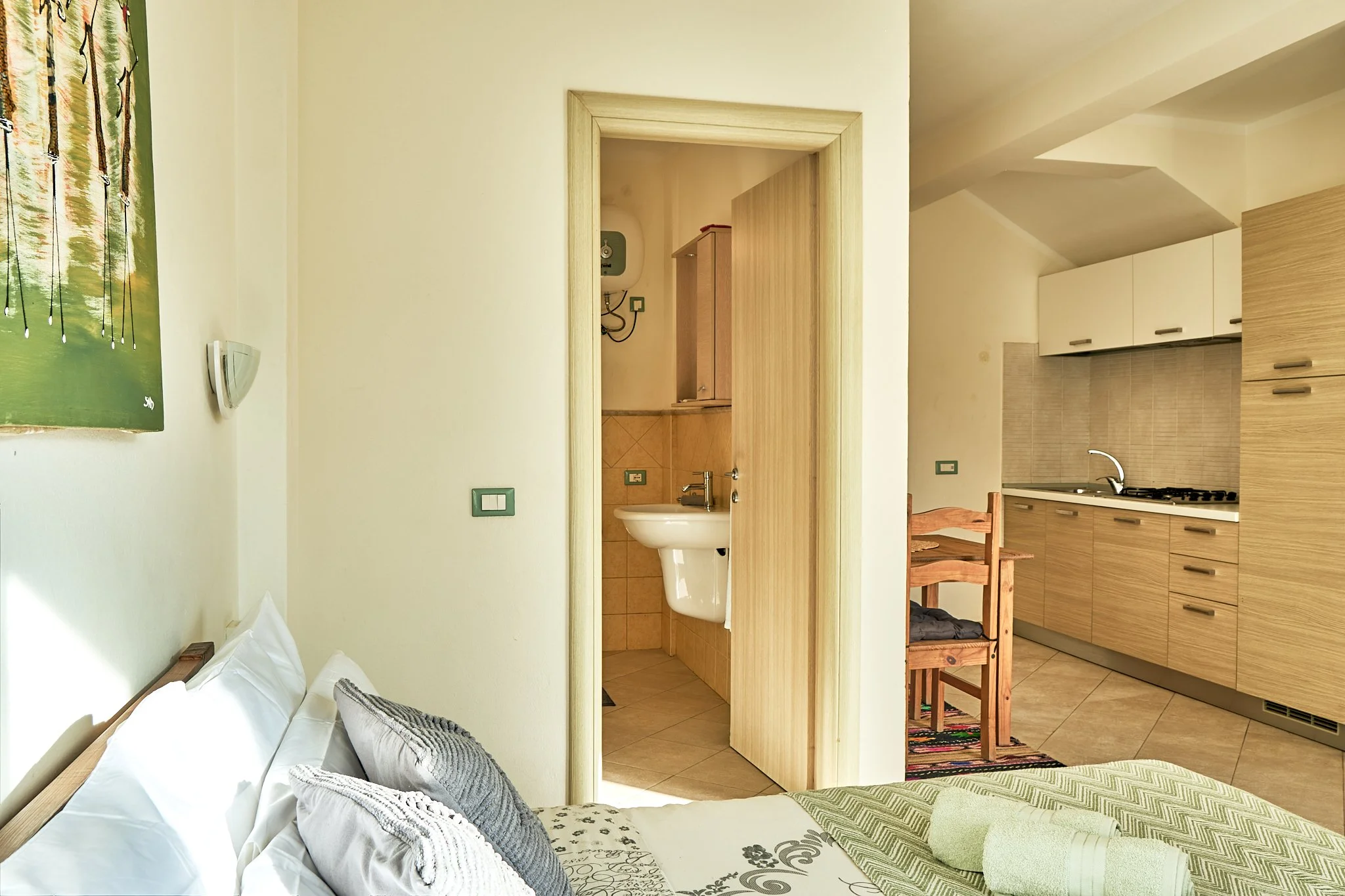 A cozy bedroom with a bed featuring white and gray pillows, a bedside table, and a window with a nature scene. An open door leads to a small bathroom with a white sink, and beyond that is a kitchen area with wooden cabinets, a countertop, and a dinin