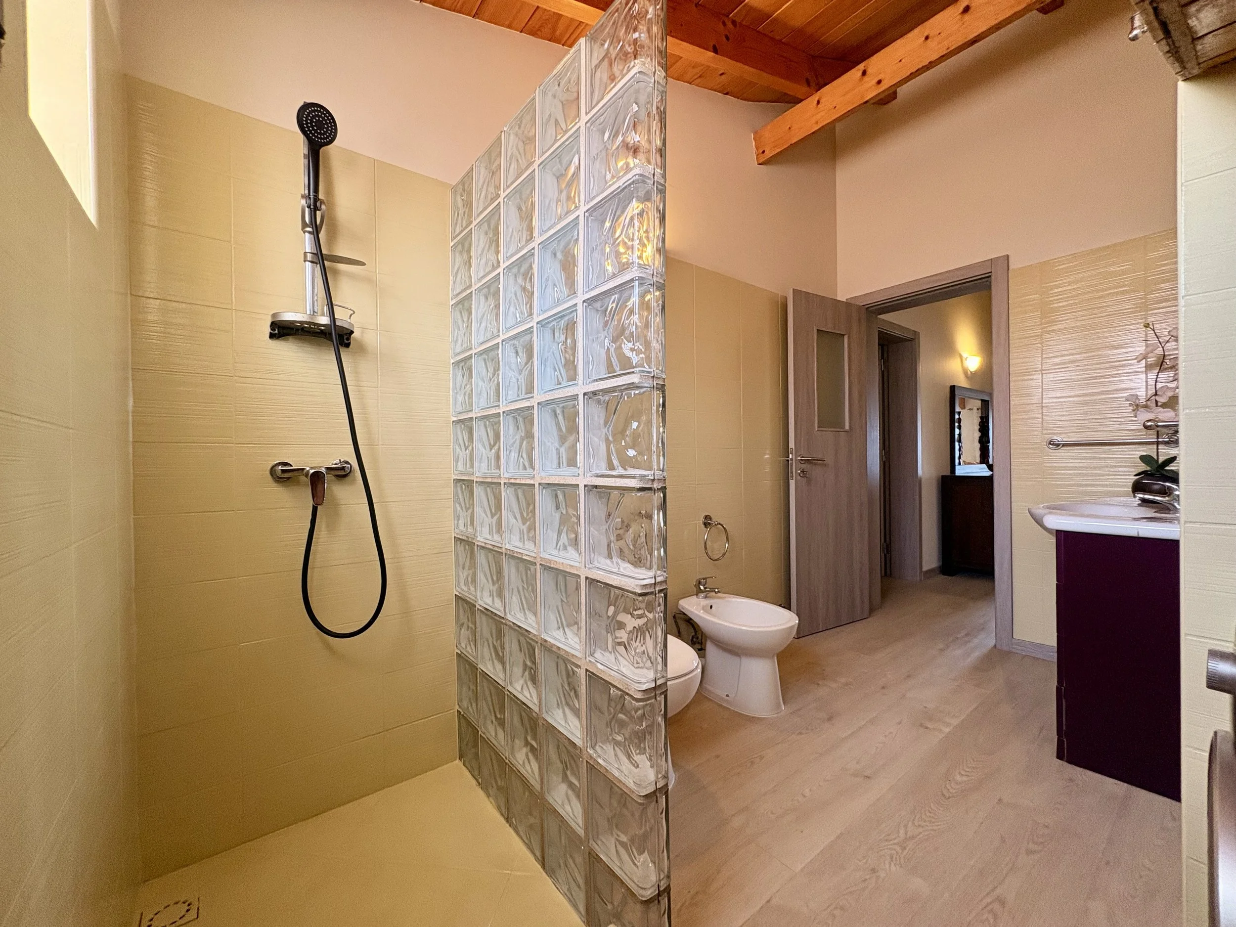 A bathroom featuring a glass block wall separating the shower area from the toilet and sink, with beige tiles and wooden ceiling beams.