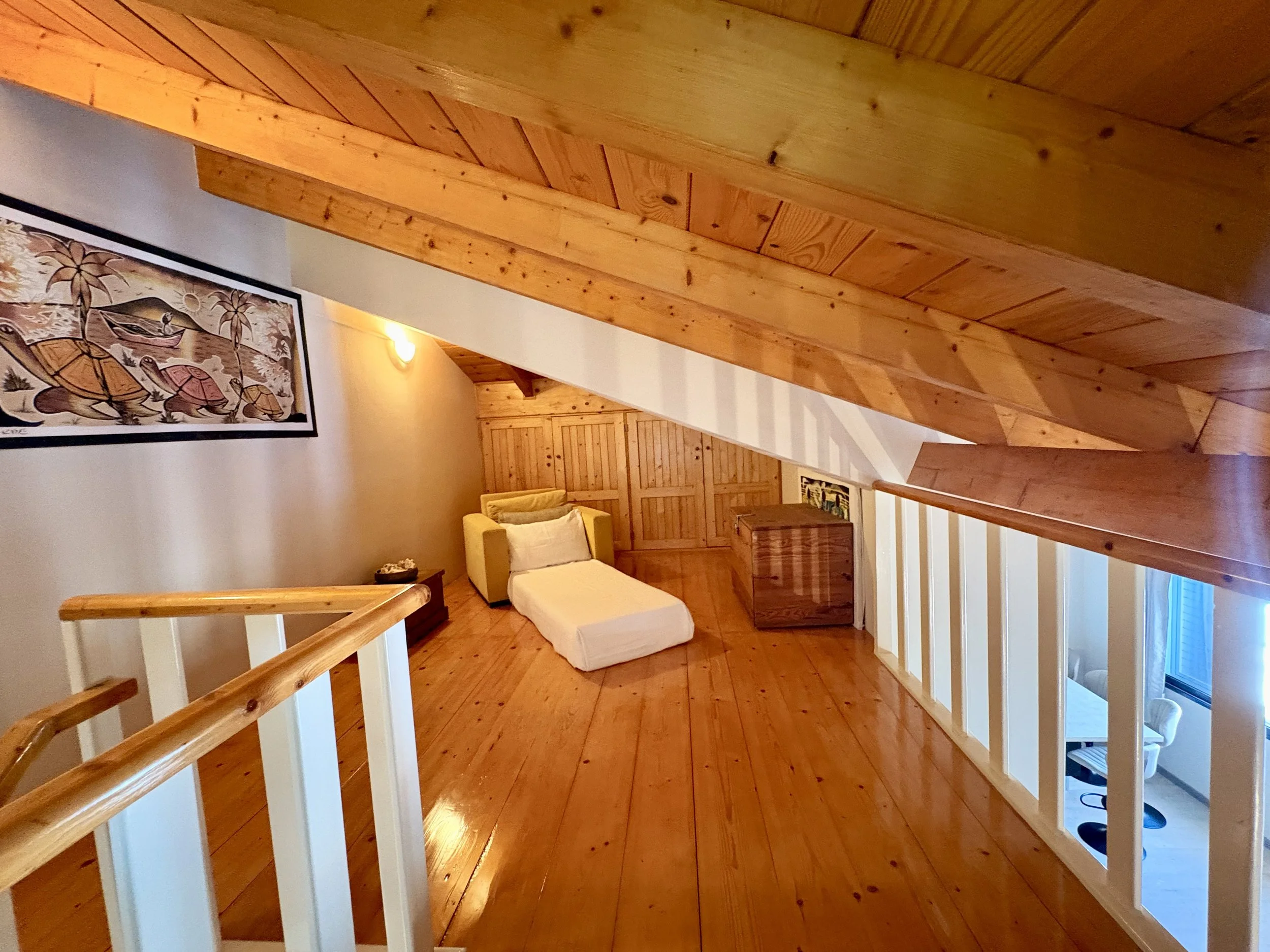 A cozy living room with wooden flooring and beams, a yellow armchair with pillows, a framed picture of an African scene on the wall, and a staircase with white railing. There is warm lighting and a wooden door in the background.