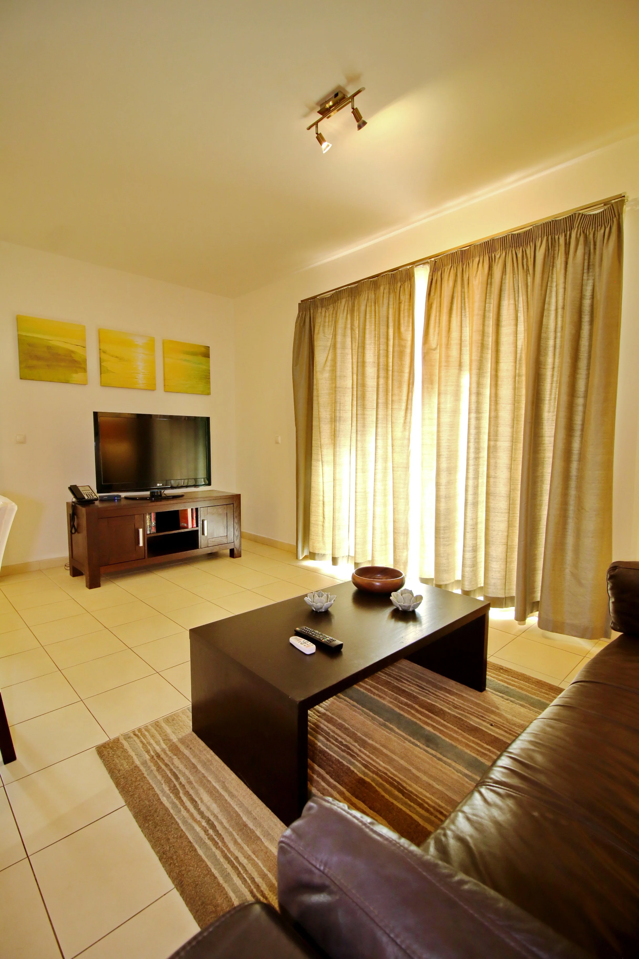 Living room with beige curtains, a dark wood coffee table, a brown leather sofa, a television on a wooden stand, framed paintings of a sunset, and a striped area rug.