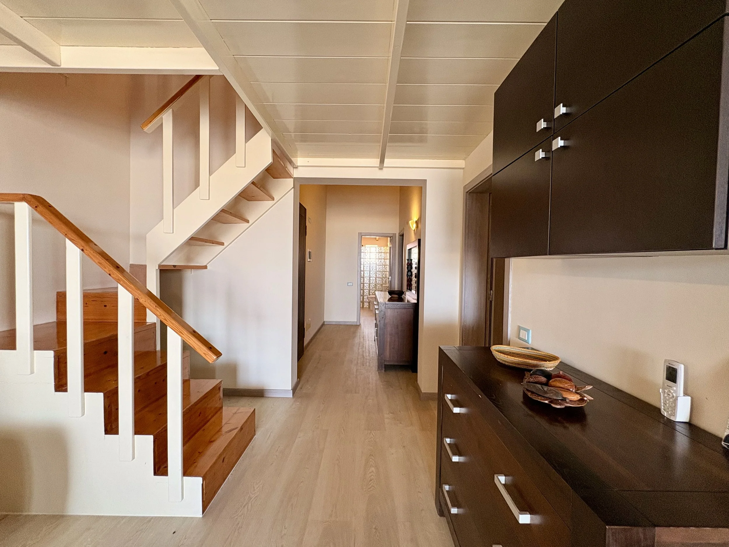 Interior of a house with a staircase, wooden flooring, dark wooden cabinet, and various decor including a bowl and remote control on a console table.
