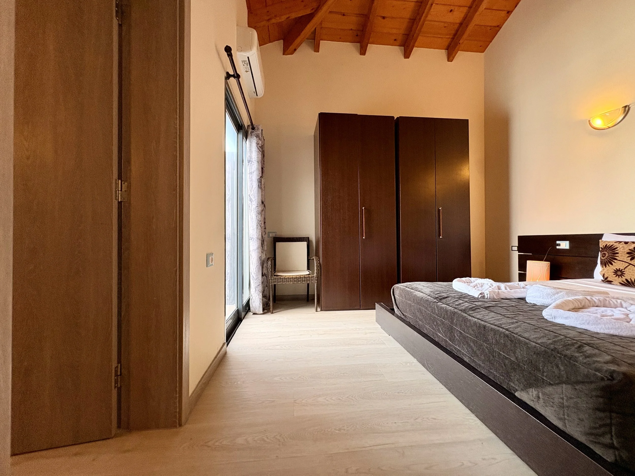 A bedroom with wooden ceiling beams, beige walls, and light wooden flooring. There is a bed with a dark gray mattress and folded white linens at one end. A wooden wardrobe is against the wall, and a sliding glass door with a curtain leads to a balcon