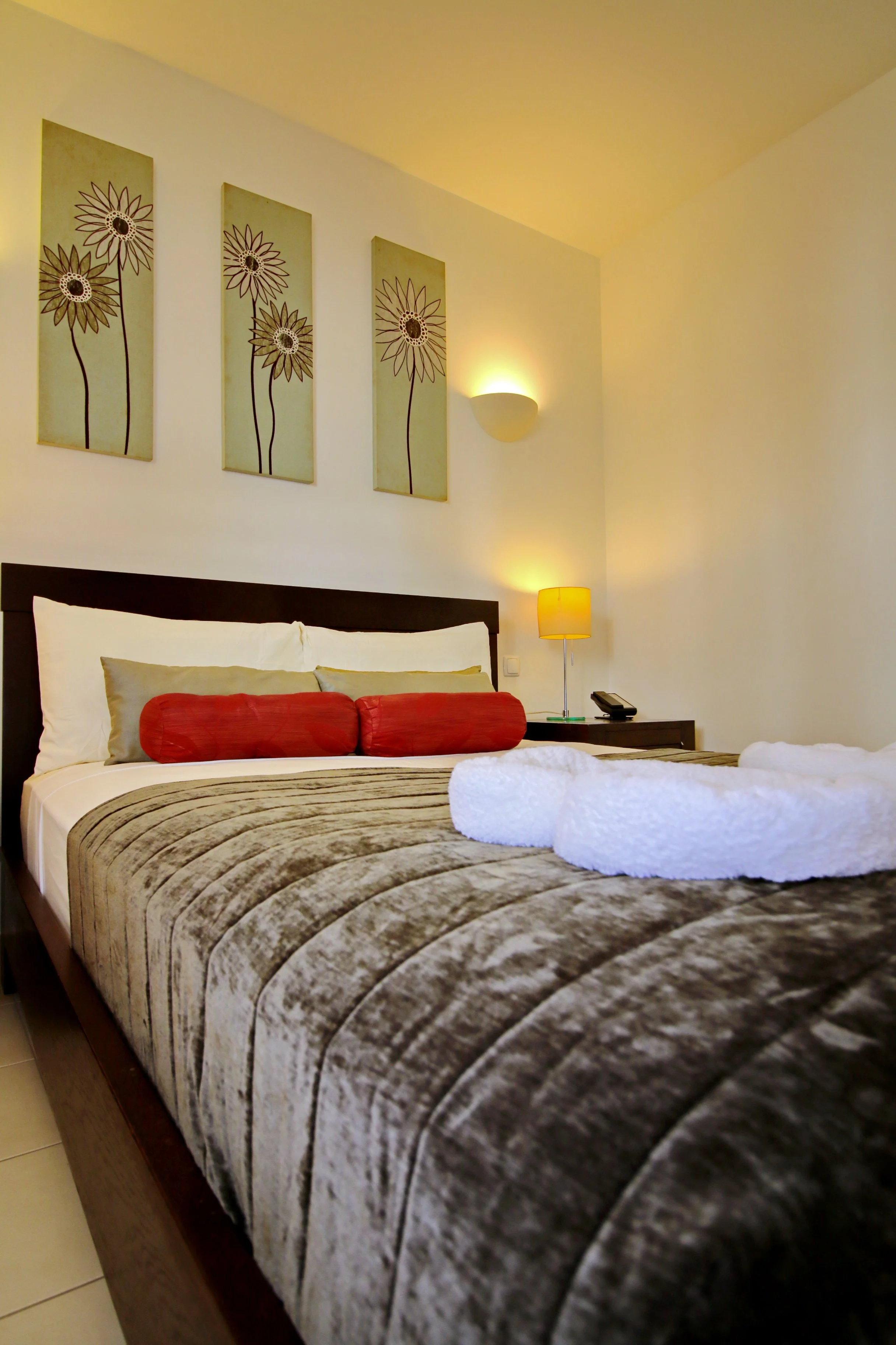 A neatly made bed with a striped bedspread, two rolled red pillows, and one long white towel arranged on top. There are three framed floral artwork pieces hanging above the bed on the wall. A wall-mounted light and a standing lamp provide warm lighti