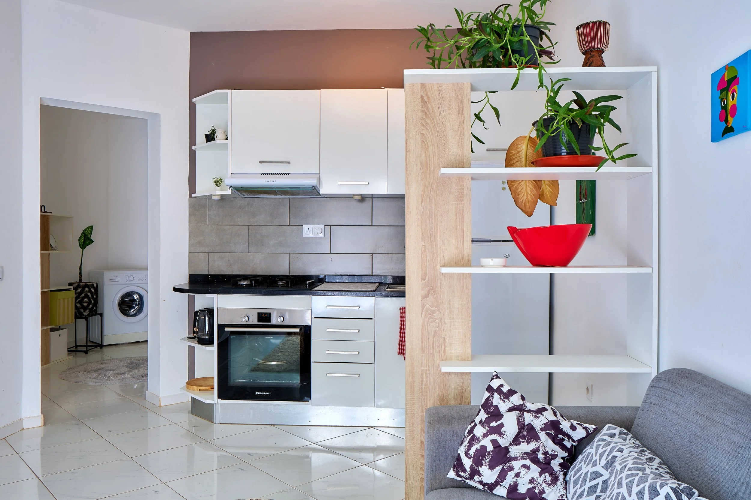 Modern kitchen with white cabinets, gray tiled backsplash, gas stove, and open shelving with potted plants and decorative items, adjacent to a living area with gray couch and patterned cushions.