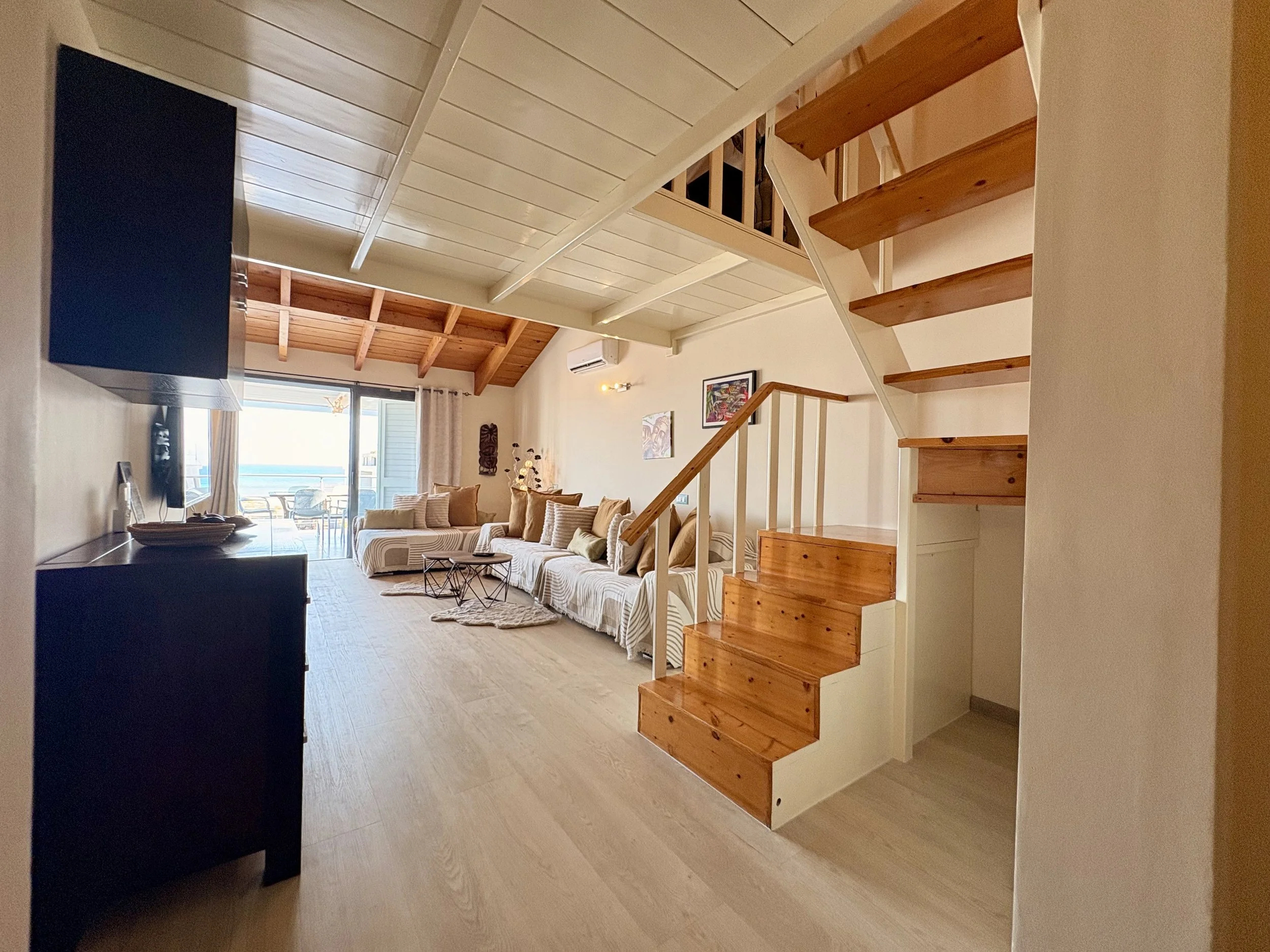 Living room with a vaulted ceiling, wooden beams, a large white sofa with multiple pillows, an art piece on the wall, a sliding glass door leading to a balcony, and a staircase with wooden steps.