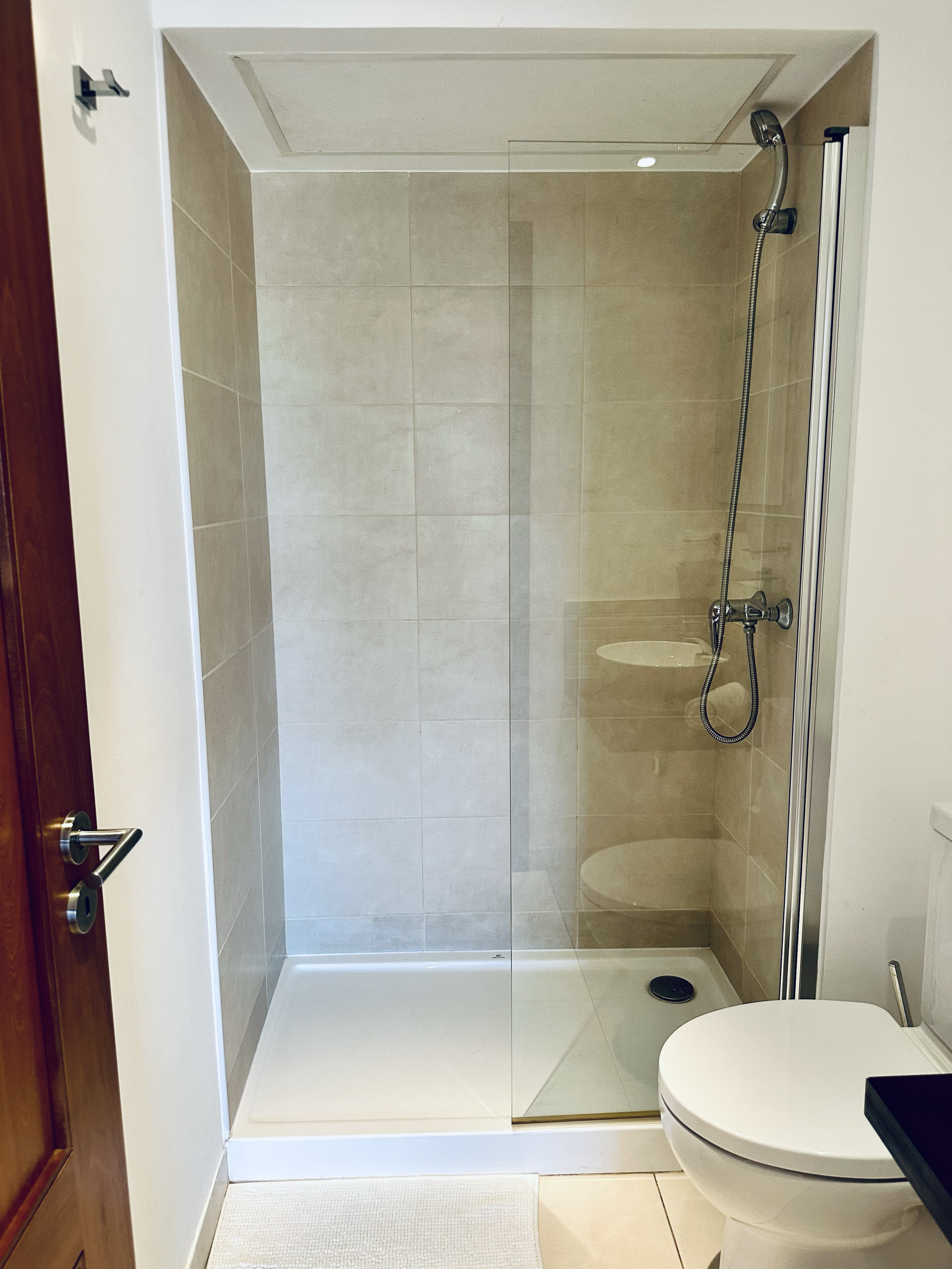 Modern bathroom with a glass shower enclosure, beige tiles, a white toilet, and a wooden door.