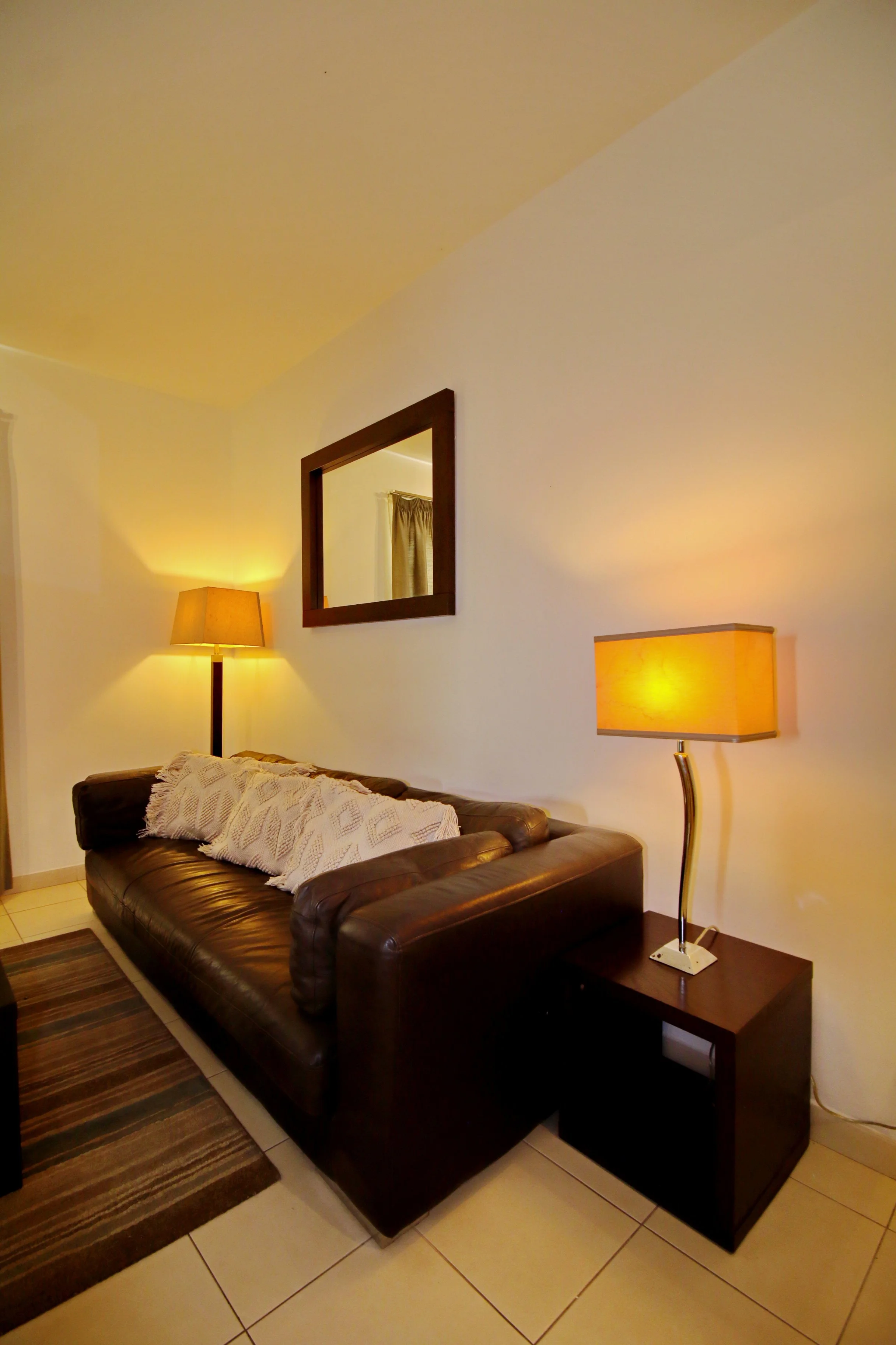 Living room with a brown leather sofa, a wooden side table with a lamp, a rectangular mirror on the wall, and a second lamp on the table, with beige curtains and tiled flooring.