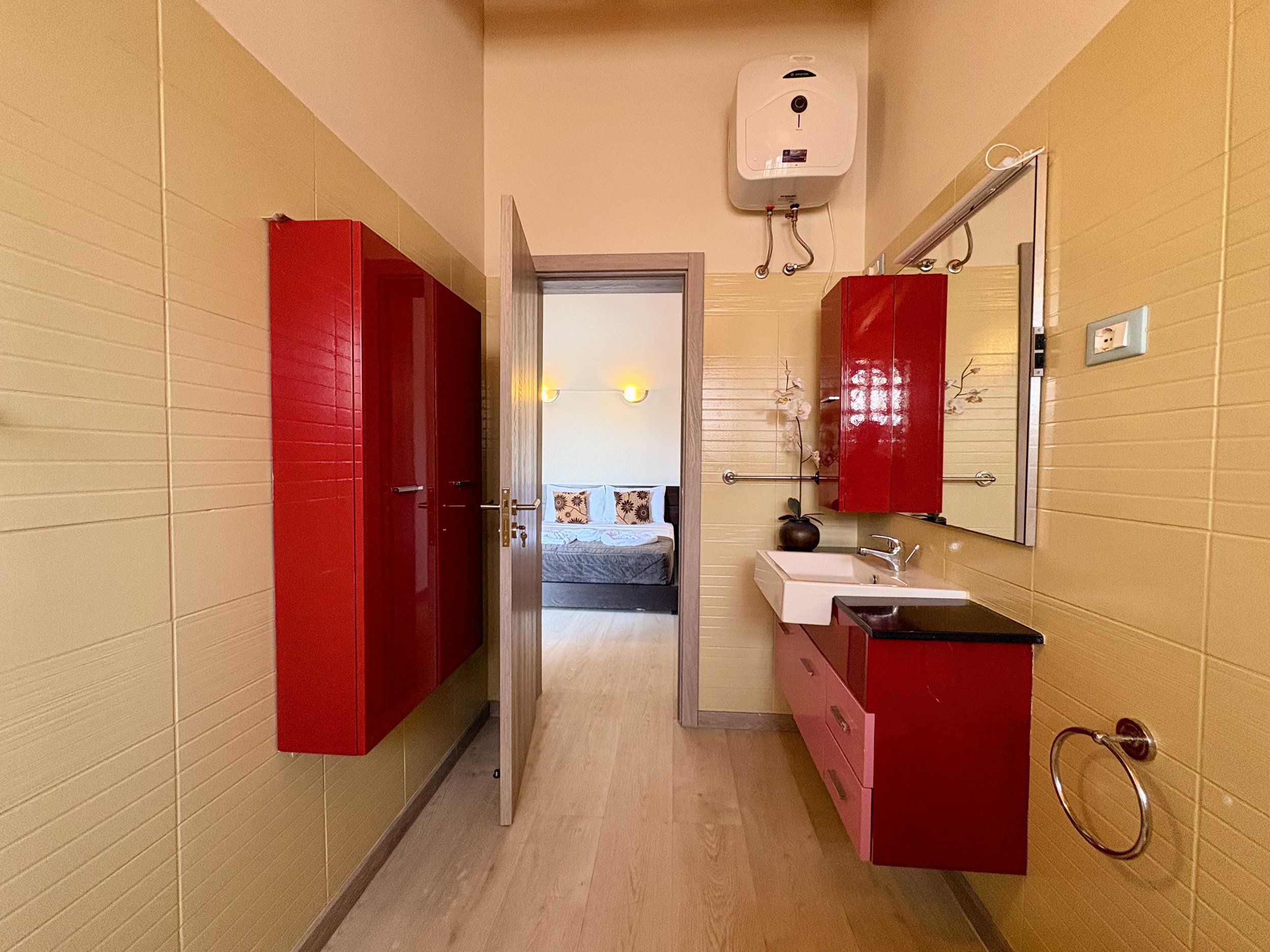 A bathroom with yellow tiled walls, red cabinets, and wooden accents. There is a white sink, a mirror, and a wall-mounted water heater. A doorway leads to a bedroom with a bed and floral pillows, visible in the background.