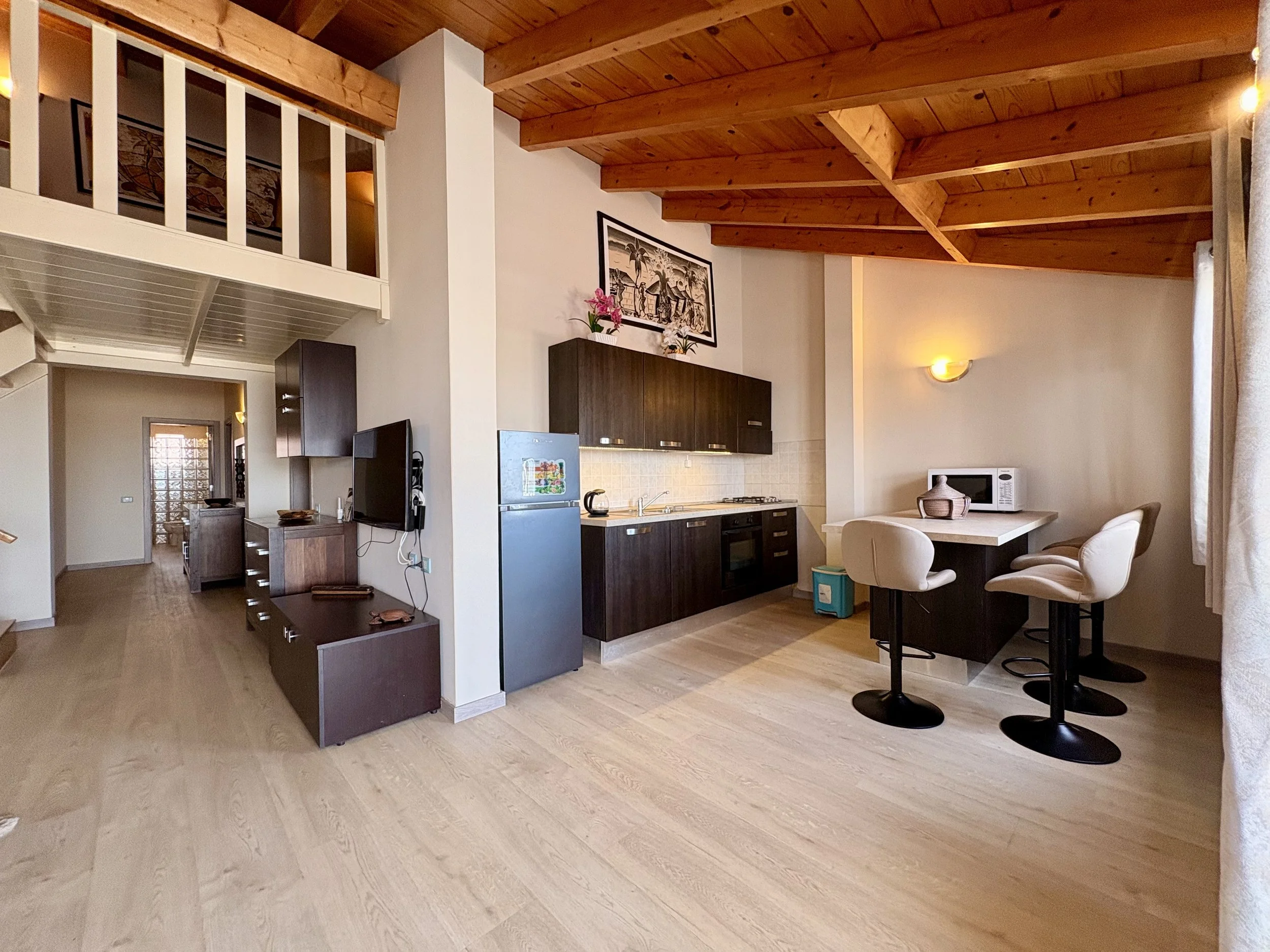 The image shows a living space with a kitchen area and a dining nook. The ceiling is made of wooden beams, and there is a whitened wall with a framed artwork. The kitchen features dark cabinets, a fridge, and some small appliances such as a microwave