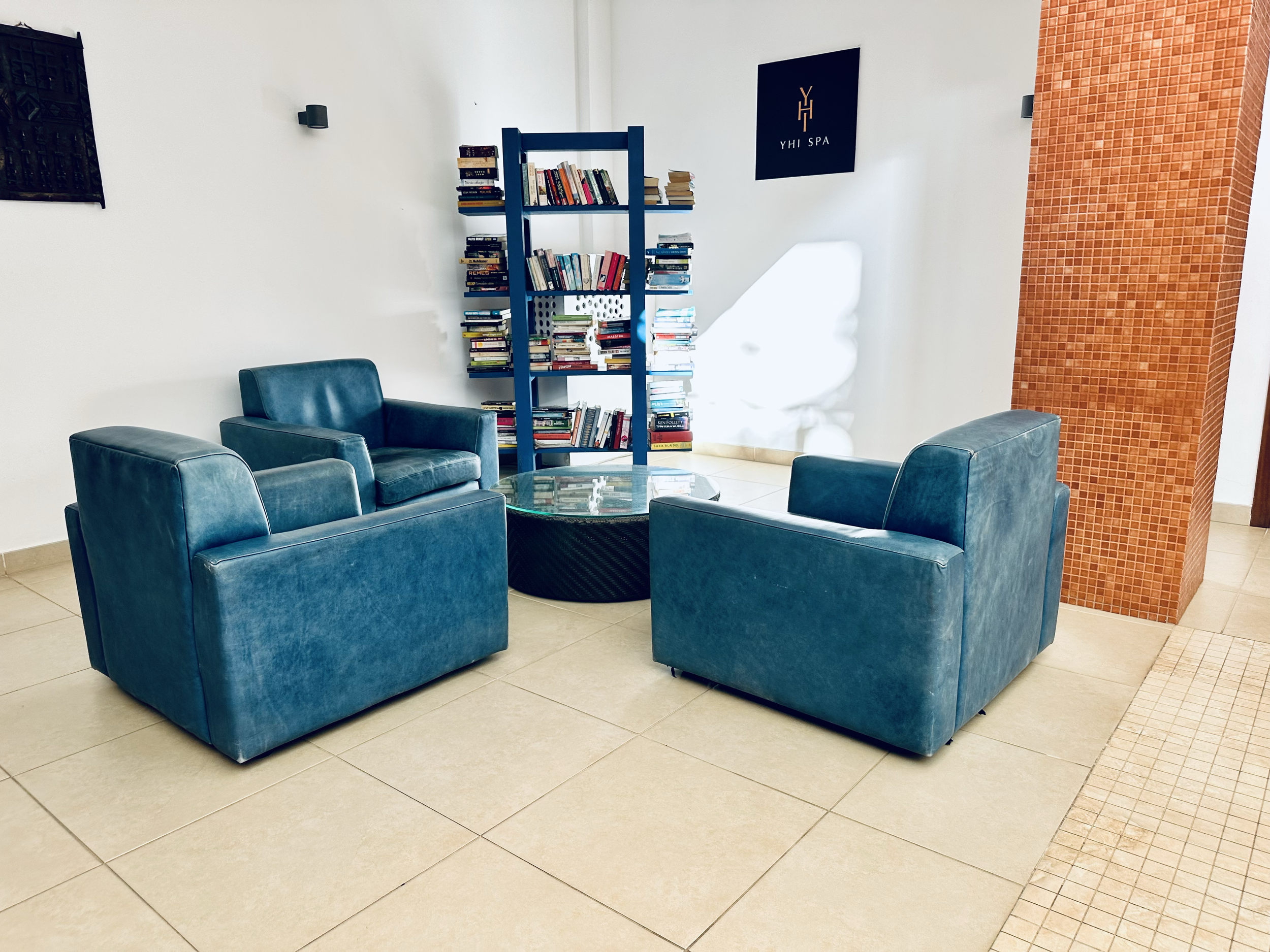 A waiting area with four teal armchairs around a glass-top coffee table, a tall blue bookshelf filled with books and stacked papers, a black sign for YHI Spa on the wall, a red mosaic-tiled column, and beige tiled floor.