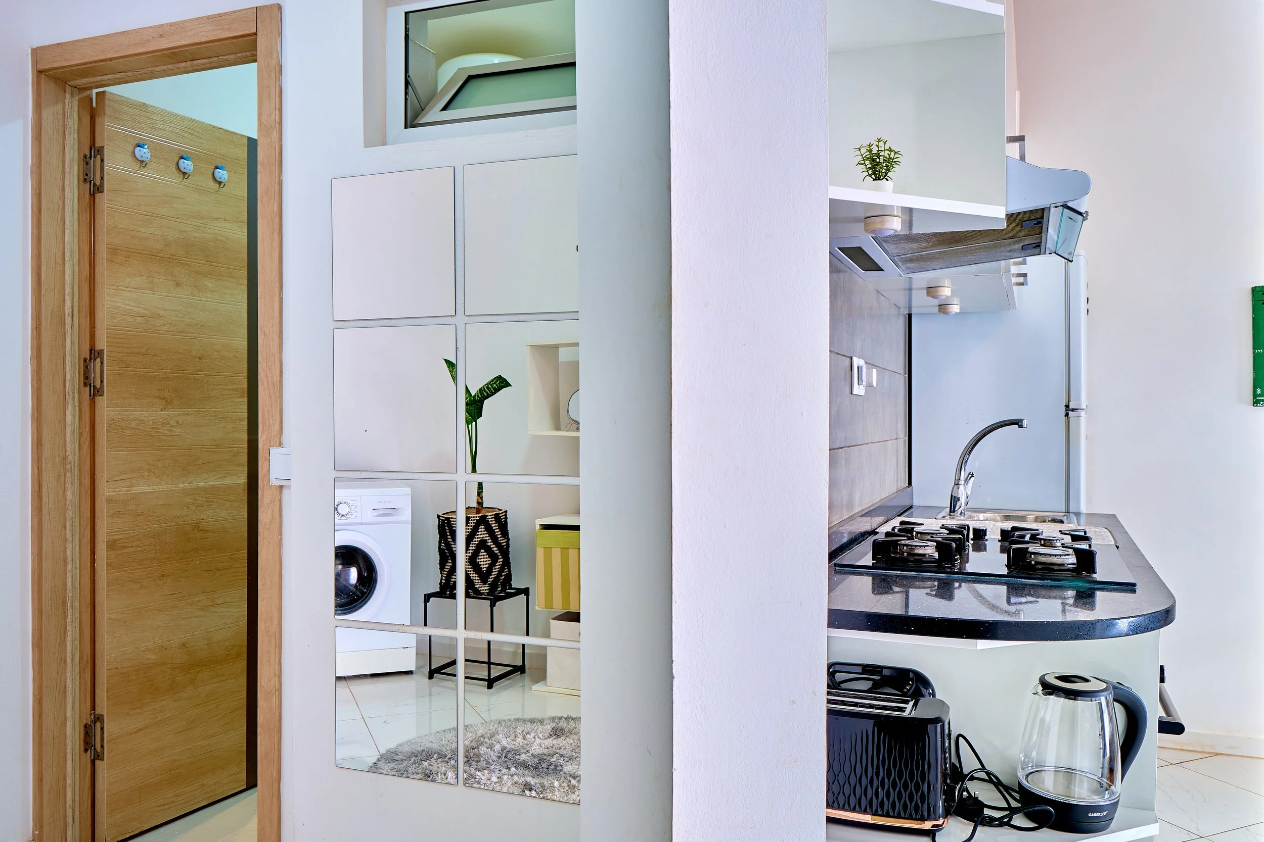 A view of a modern kitchen with a white countertop, gas stove, and a glass kettle. A mirror divides the space, reflecting a room with a washing machine, a black chair, and a potted plant. There's a wooden door frame and a small window above the count