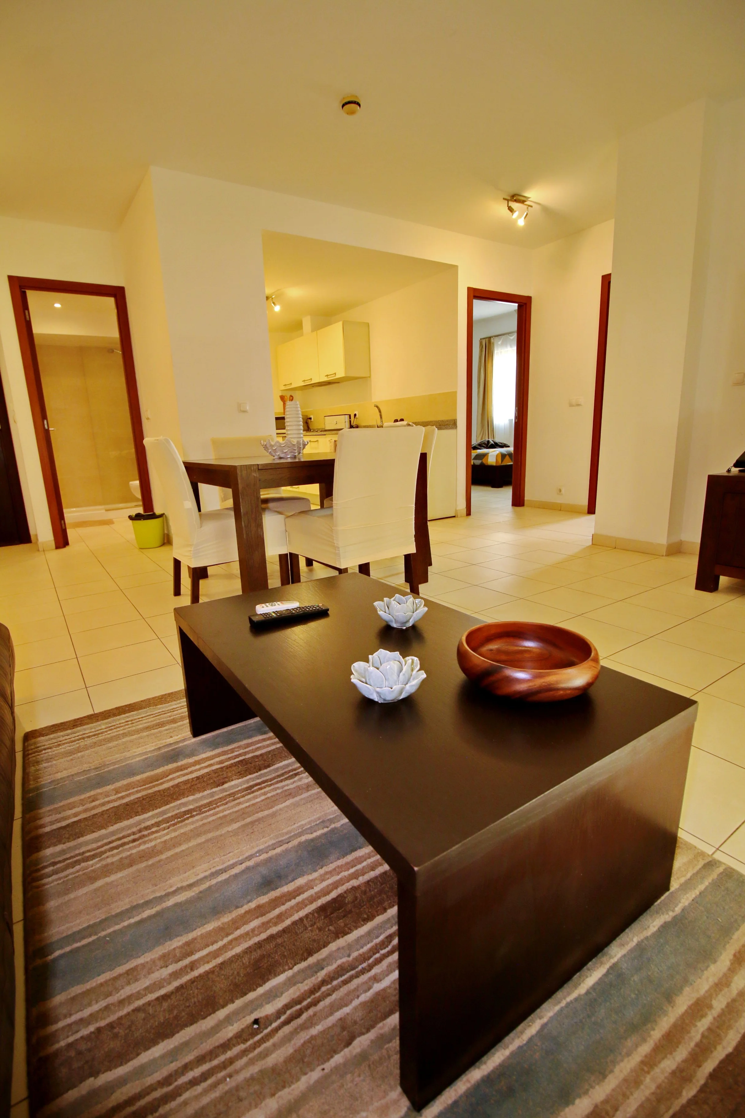 Living room with dining table, chairs, and a modern kitchen in the background. Decor includes a wooden bowl, ceramic flowers, and a striped rug.