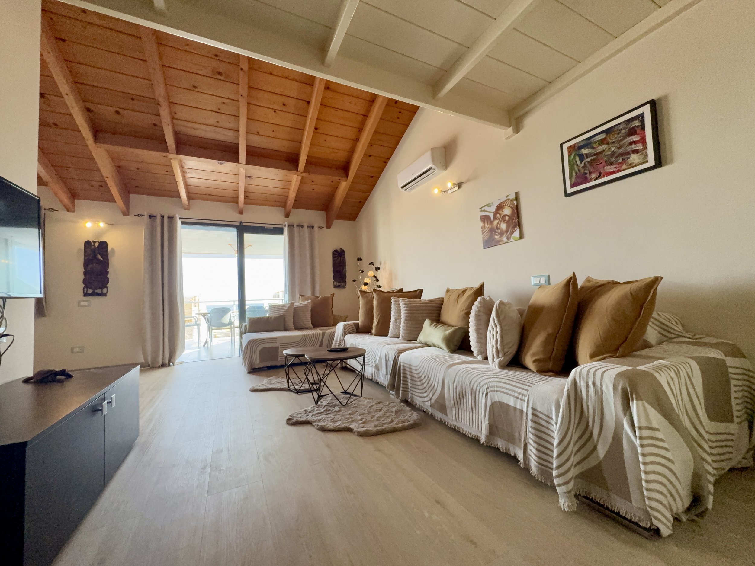A living room with a wooden ceiling, beige walls decorated with artwork, a beige couch with striped cushions, a sliding glass door leading to a balcony, and a television on the left side of the image.