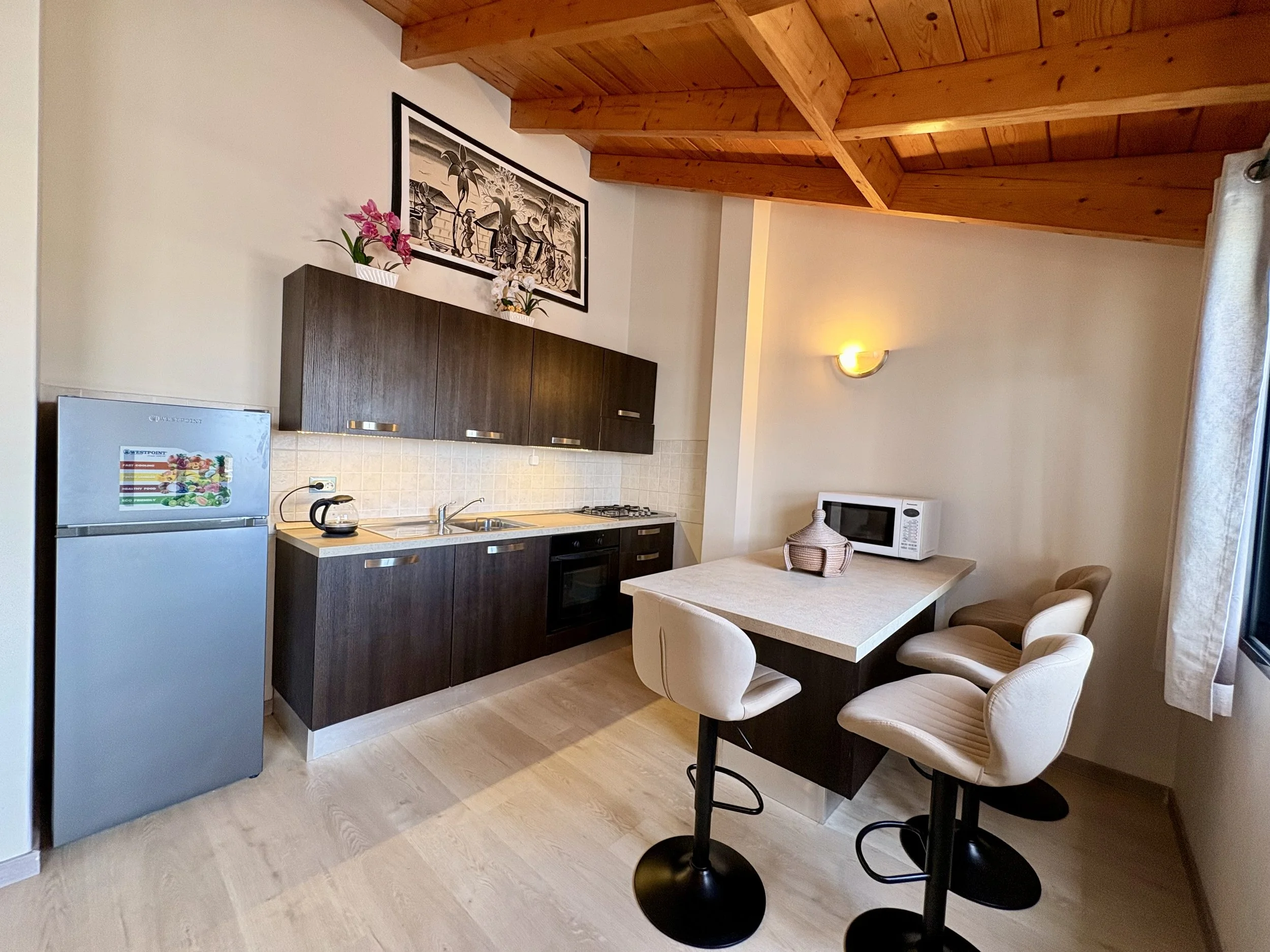 Kitchen with dark cabinets, a small refrigerator, a microwave, a table with chairs, and a wood ceiling with exposed beams.