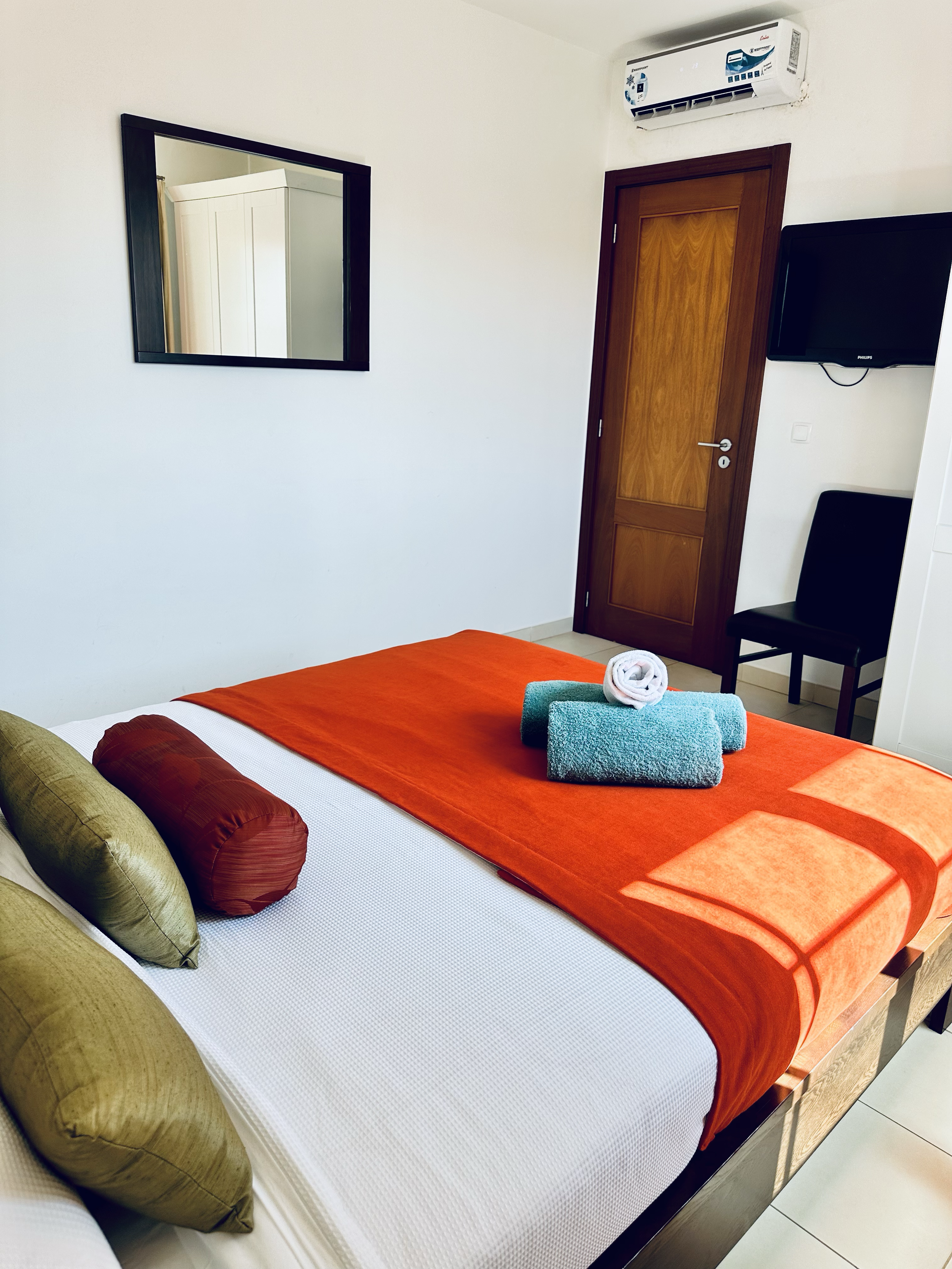A bedroom with two beds covered with white sheets and orange bedspreads, decorated with green and red pillows, and towels placed on the bed. There is a mirror, a wooden door, a wall-mounted TV, and an air conditioning unit in the room.