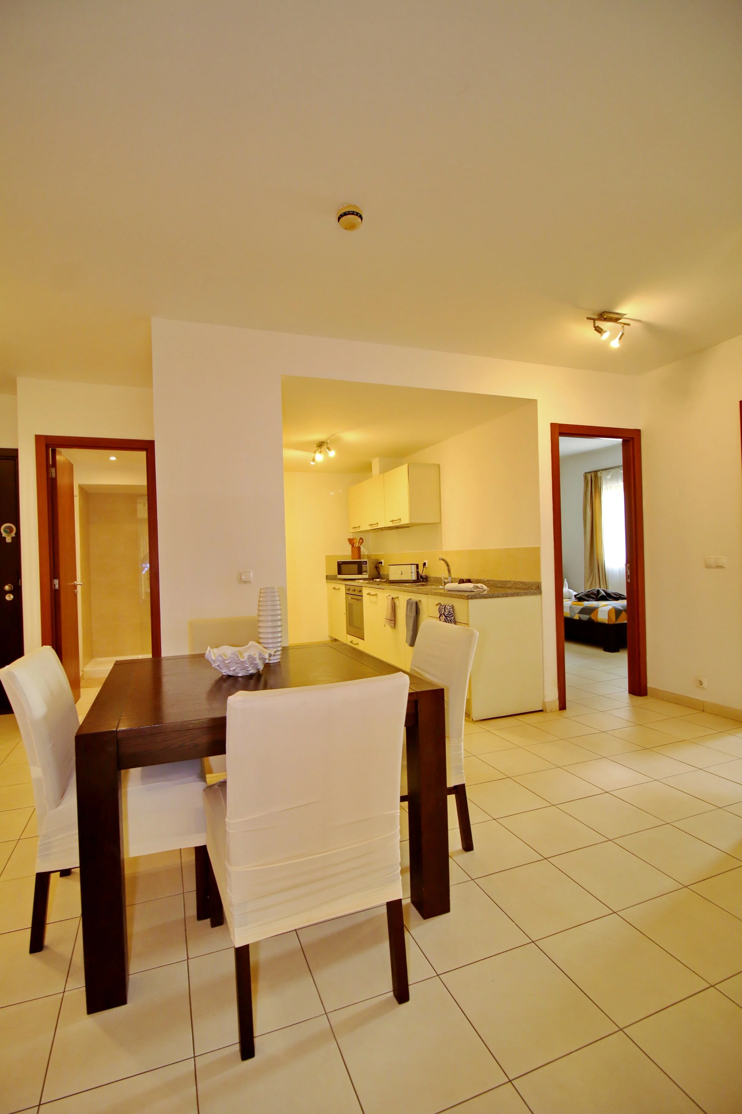 Interior view of a dining area with a dark wooden table and white chairs, and a kitchen in the background with white cabinets and beige tiled floors.