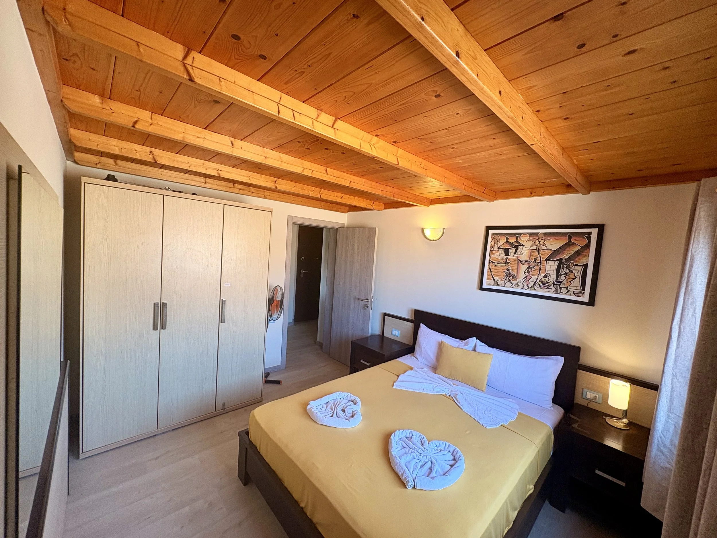 A bedroom with a wooden ceiling, a bed with yellow bedding, towel art on the bed, a large closet, a nightstand with a lamp, a framed artwork on the wall, and an open door.