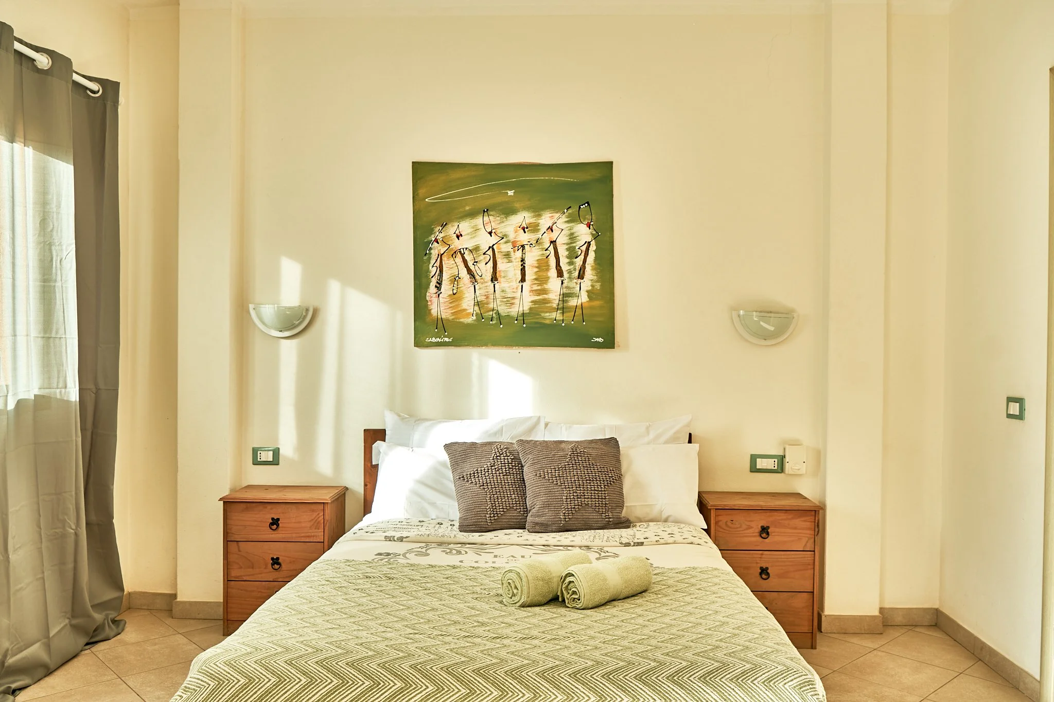 A bedroom with two wooden nightstands, a bed with pillows and rolled towels, wall art above, and curtains on the left side.