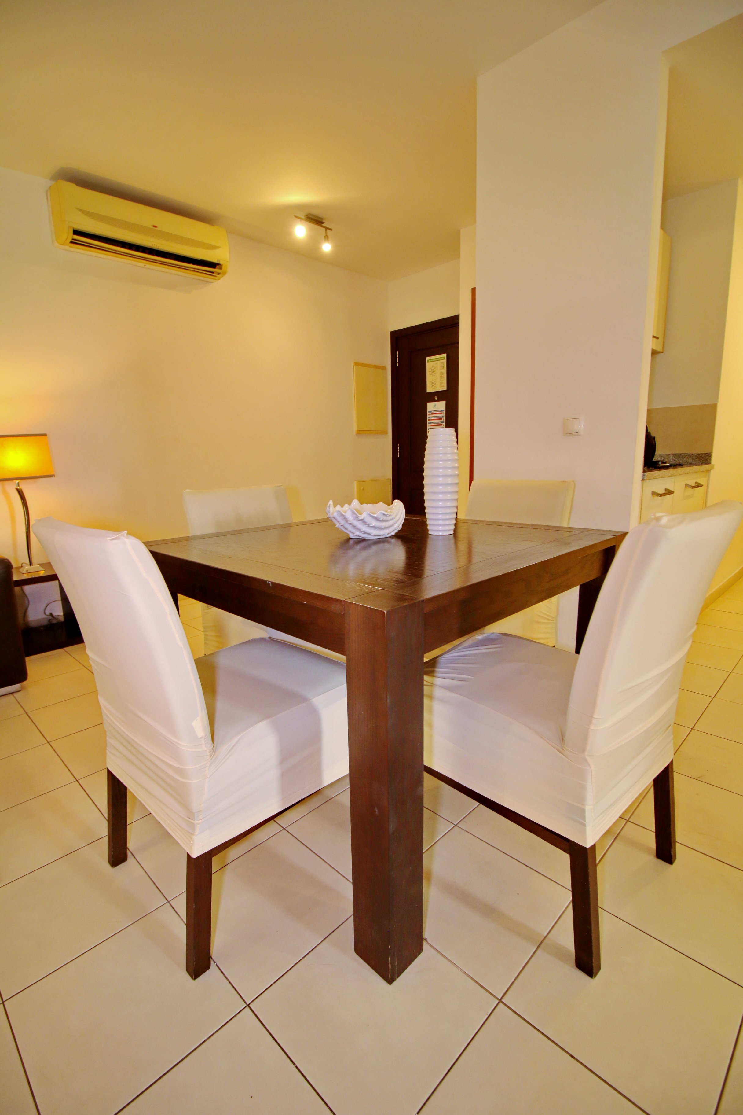 Dining room with a dark wood table surrounded by white upholstered chairs, a beige tiled floor, a small lamp on a side table, and decorative vases on the table, with an air conditioning unit on the wall and a doorway leading to another room.