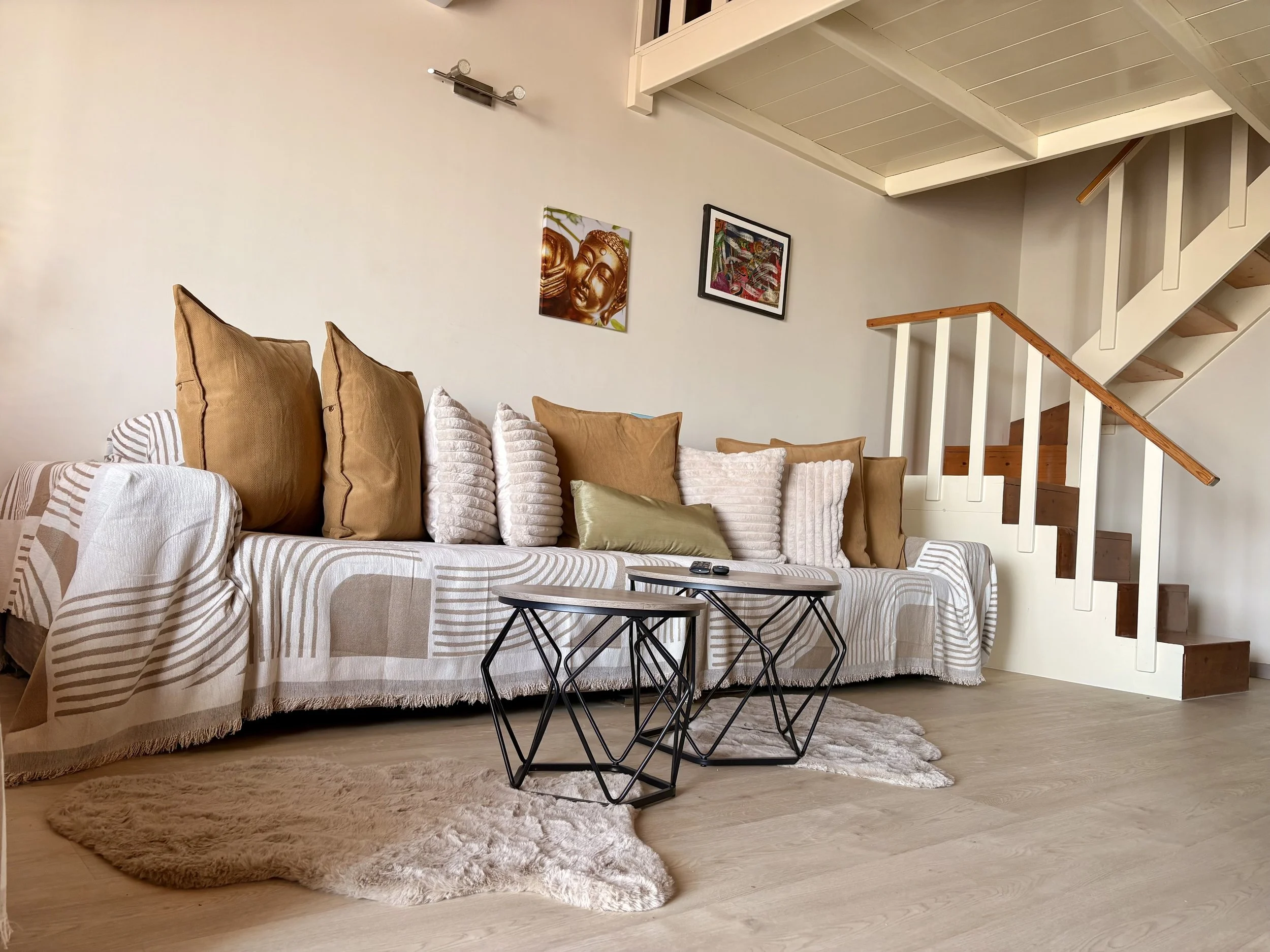 Living room with beige cushioned sofa, various pillows, black coffee tables, a plush beige rug, wall art, and a staircase with white railing and wooden steps.