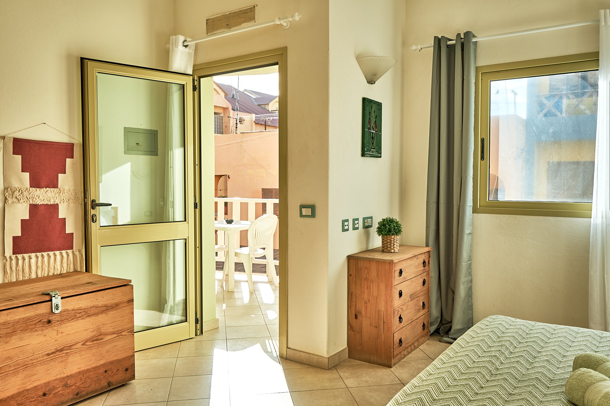 Interior of a bedroom with a wooden dresser and a bed, opening to a balcony with white chairs and a view of neighboring buildings and rooftops.