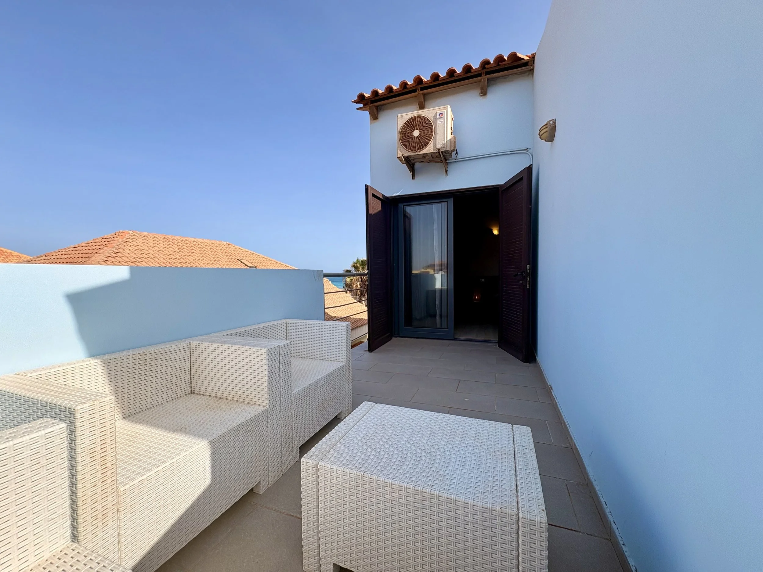 Rooftop terrace with white wicker furniture, open door to indoor space, blue wall, and clear sky in the background.