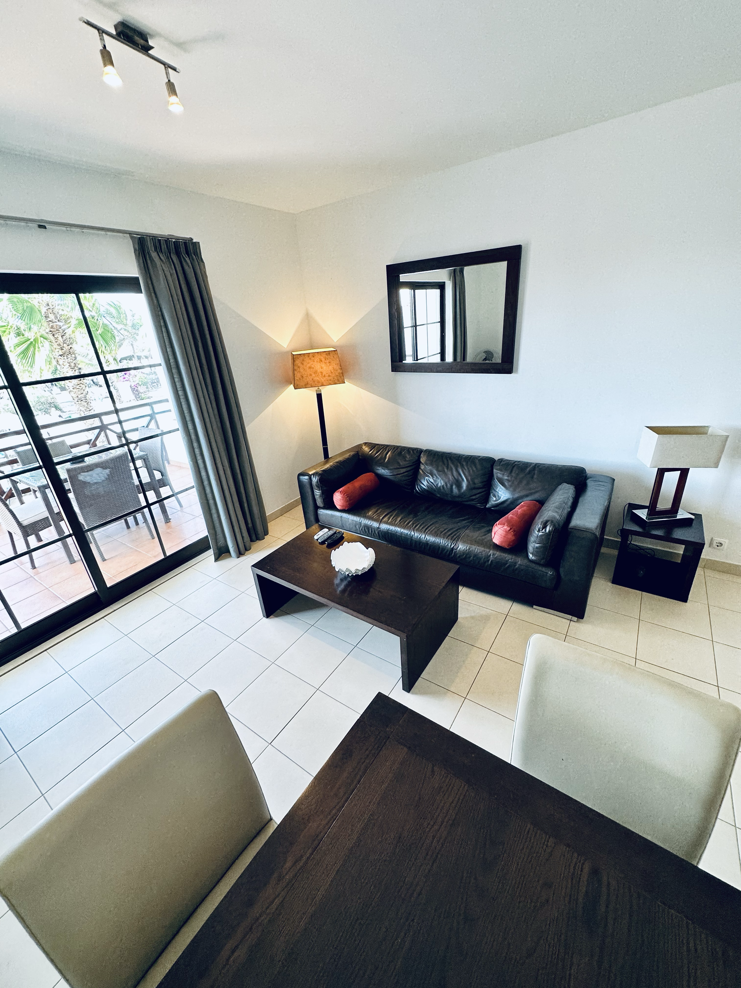 Living room with black leather sofa, side table with lamp, window with dark curtains, and a balcony with outdoor seating.