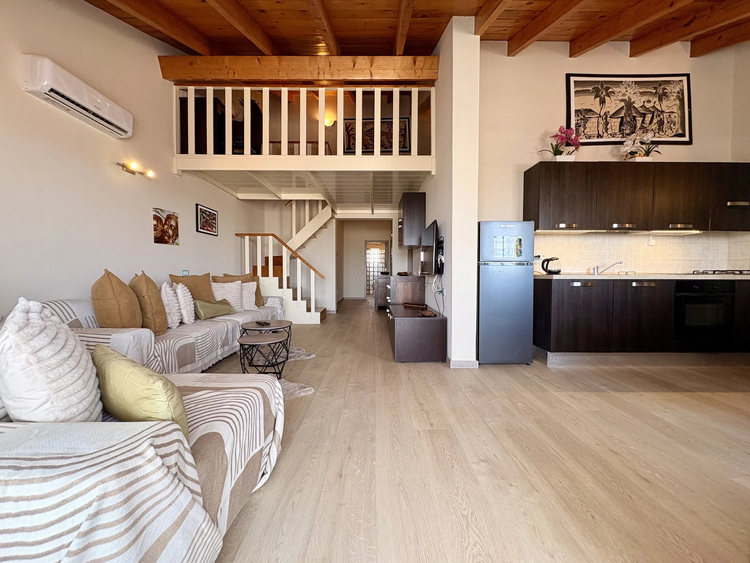 Living room with a sofa, cushions, and a coffee table, overlooking a staircase and a loft area with wooden ceiling beams, a kitchen with dark cabinets, a refrigerator, and wall art with plants.