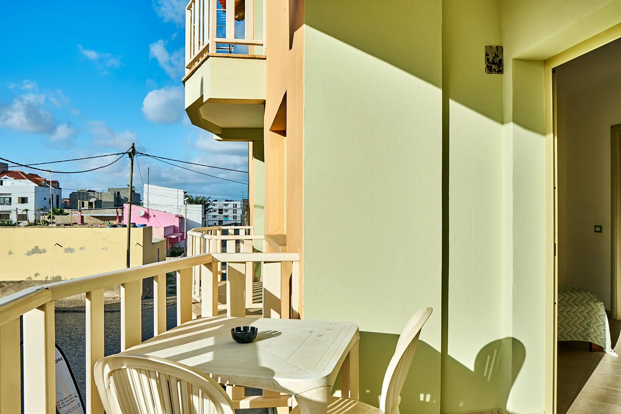 Balcony with cream-colored plastic table and chairs, green exterior wall, with city buildings and blue sky in the background.