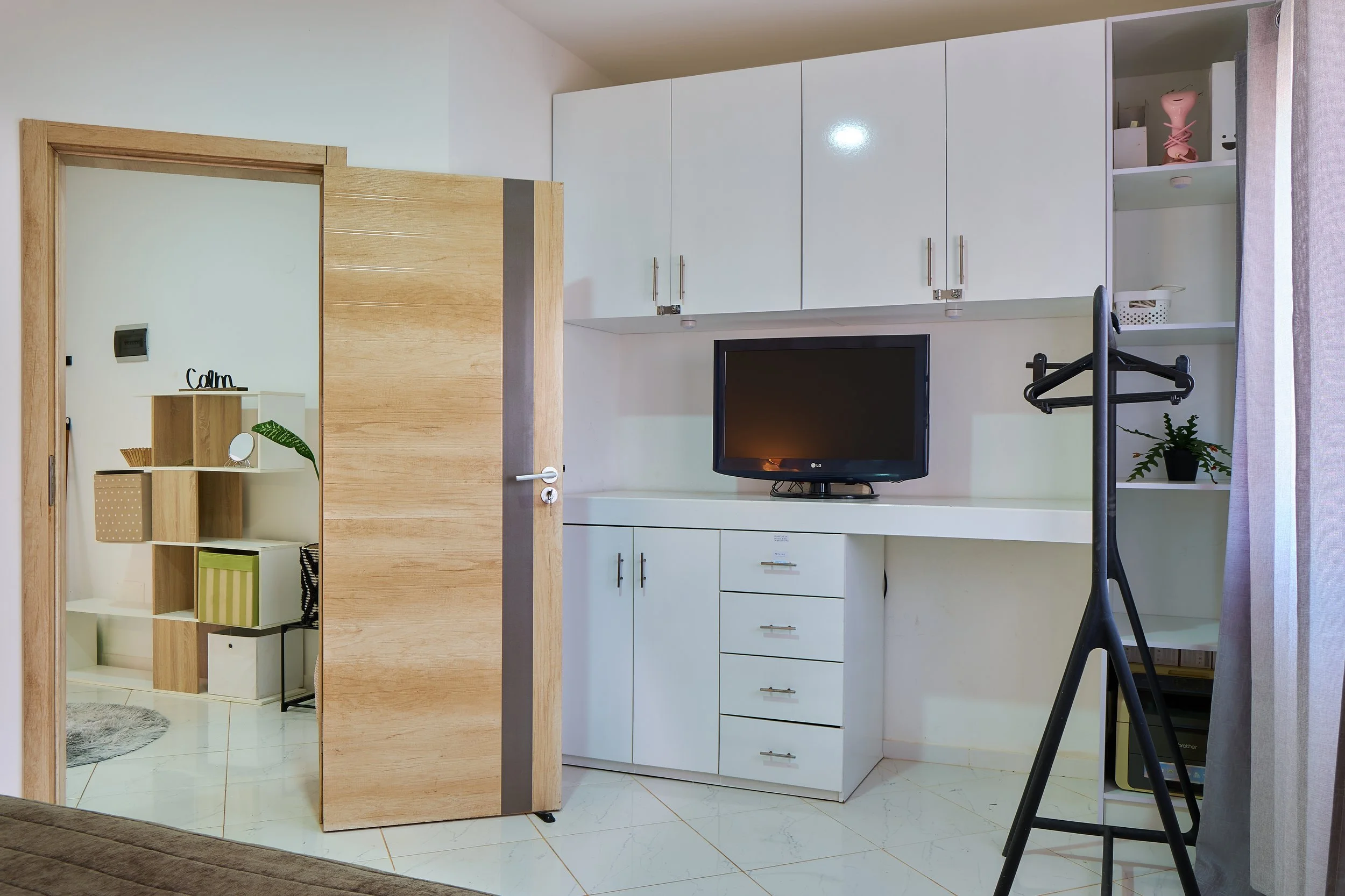 Interior of a room with white cabinets and a television, seen through an open wooden door, with a small shelf and decor in the adjacent room.