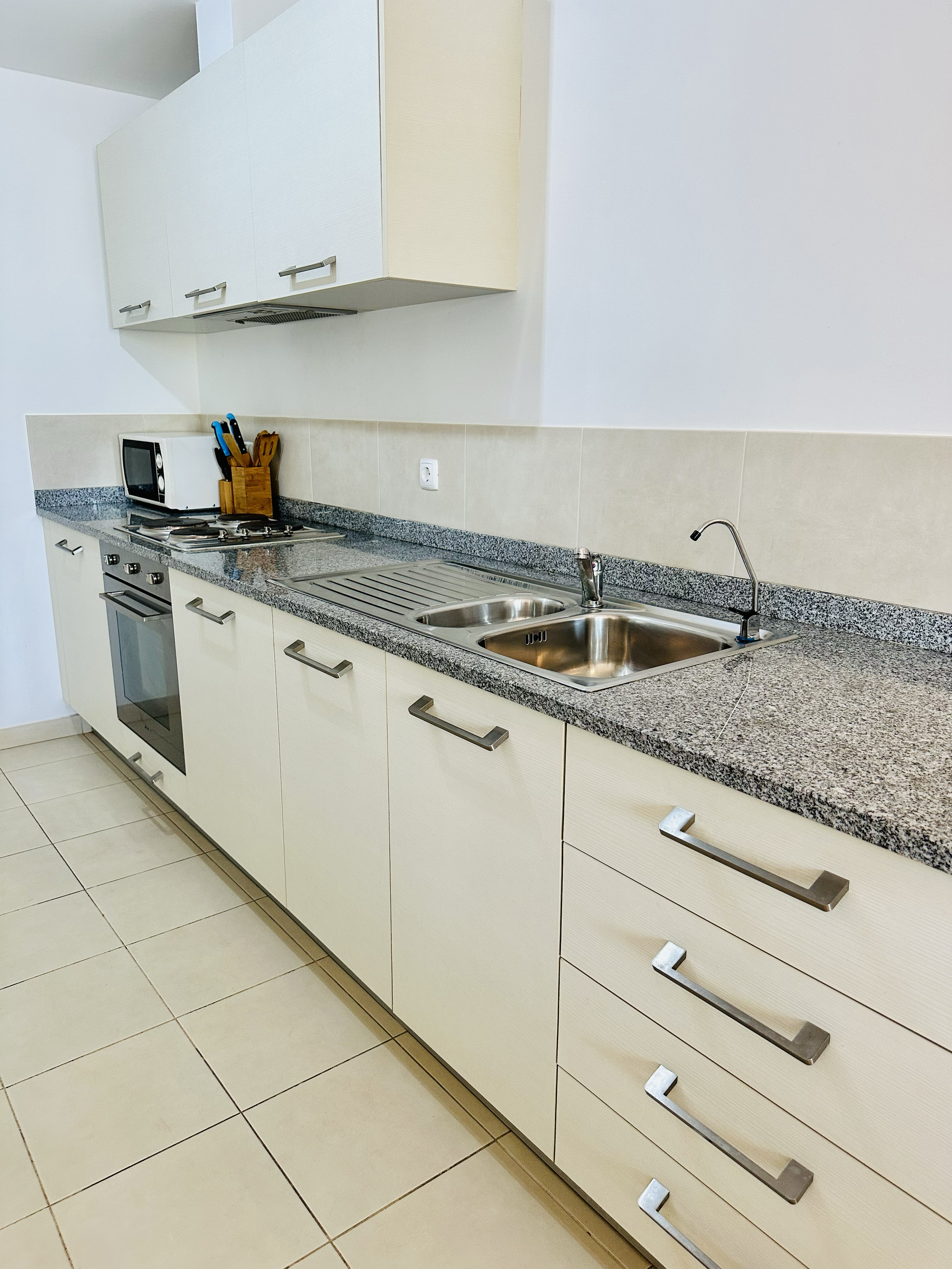 Kitchen with cream-colored cabinets, granite countertop, stainless steel sink, stove, microwave, utensil holder, and tiled floor.