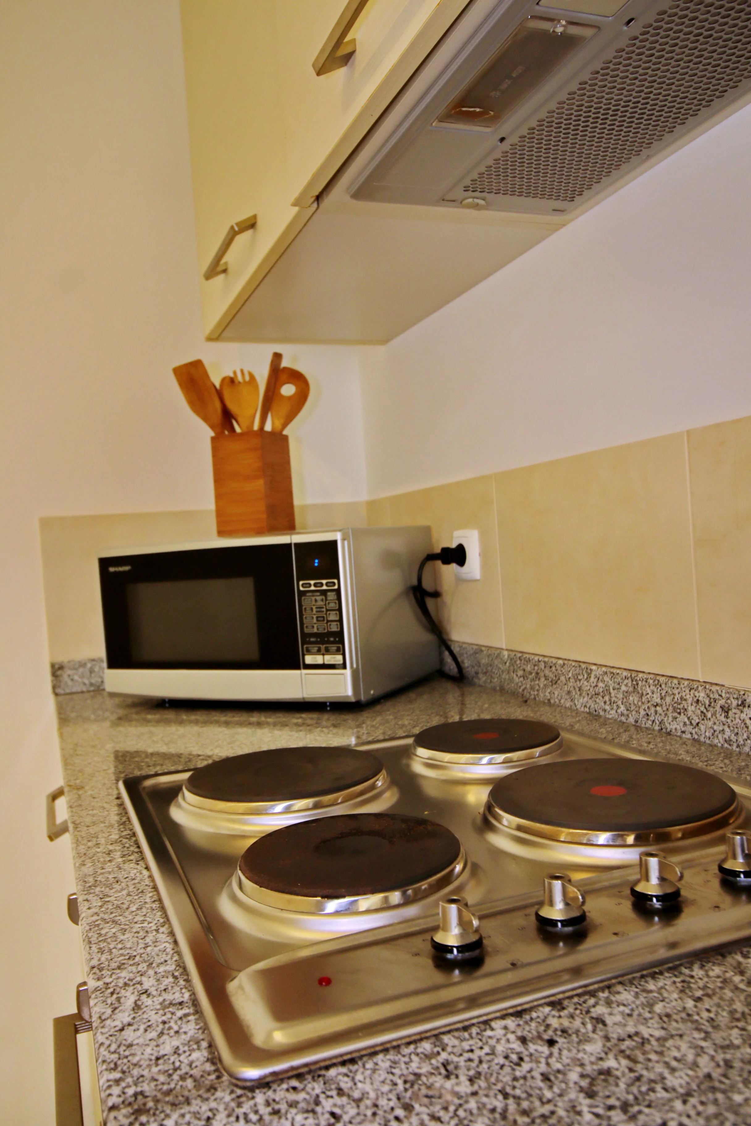 Kitchen stove with four electric burners, a microwave oven, and a wooden container holding cooking utensils on a granite countertop.