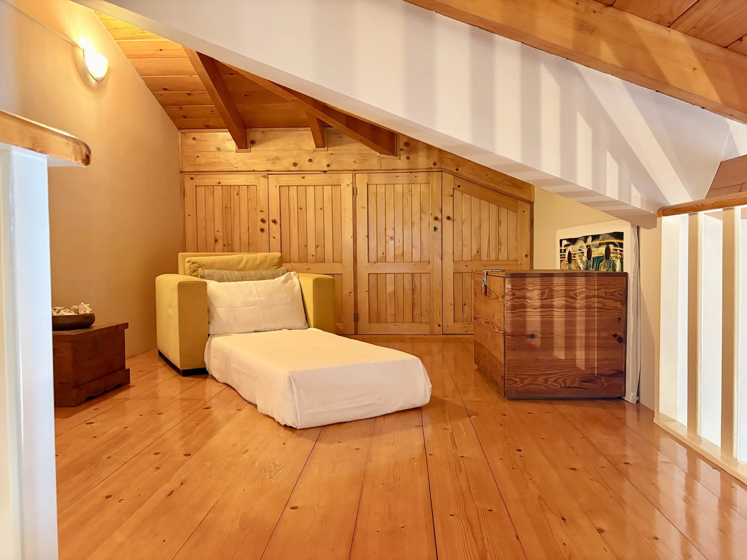 Interior view of a cozy room featuring wooden walls, ceiling, and furniture, with a yellow armchair, a small wooden side table, and a colorful piece of artwork on the wall.