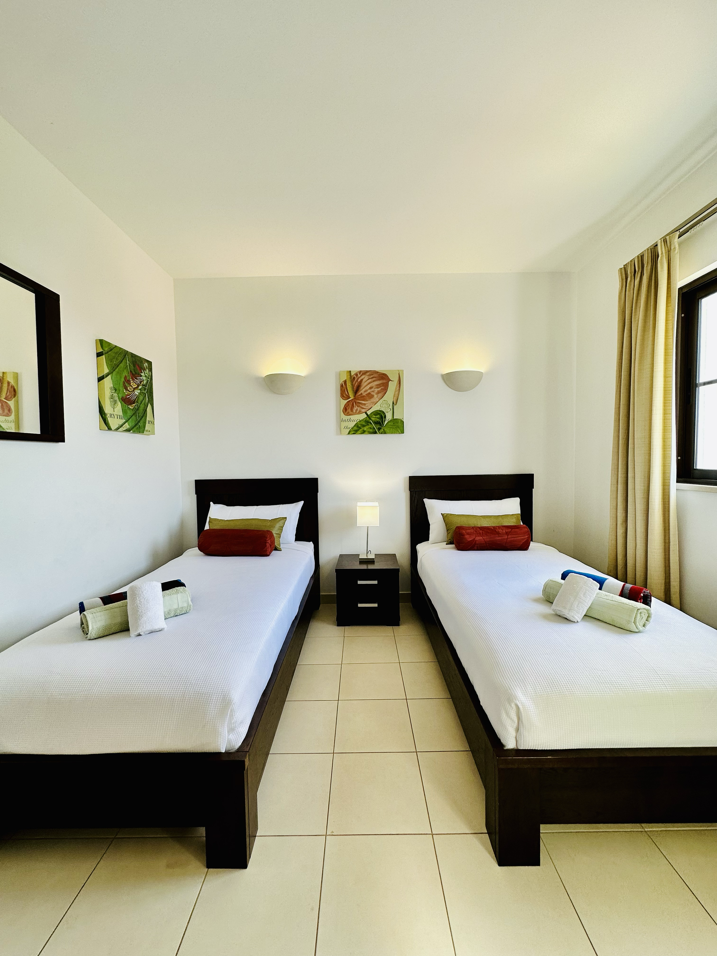 A hotel room with two single beds, white bedding, and rolled towels on each bed, placed on the foot of the beds. There is a small dark brown nightstand between the beds with a table lamp. The wall above has two wall-mounted lights and a floral painti