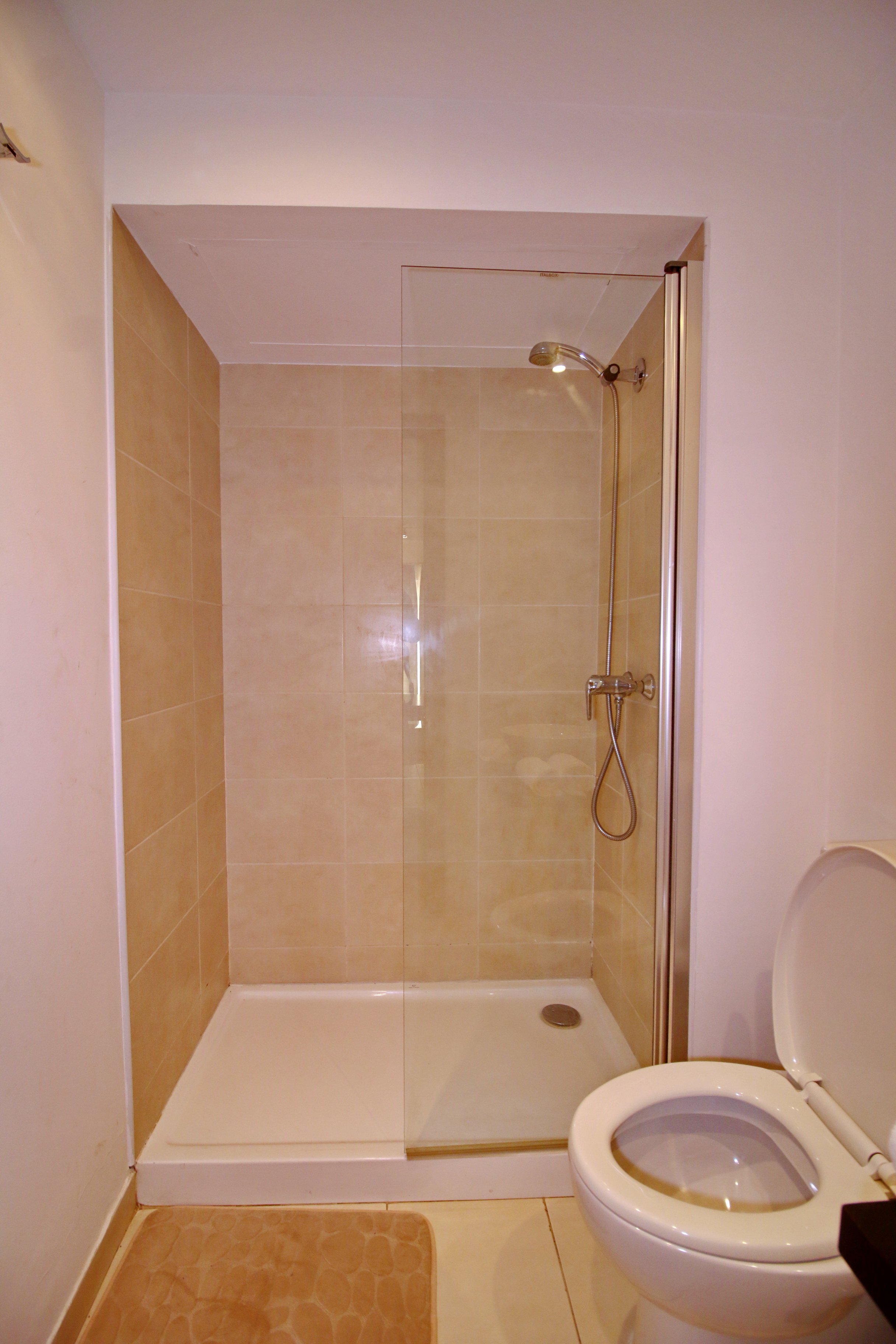 A bathroom with a walk-in shower featuring beige tiles, a handheld showerhead, a drain, and a white toilet with the lid open. There is a beige shower mat on the floor outside the shower.