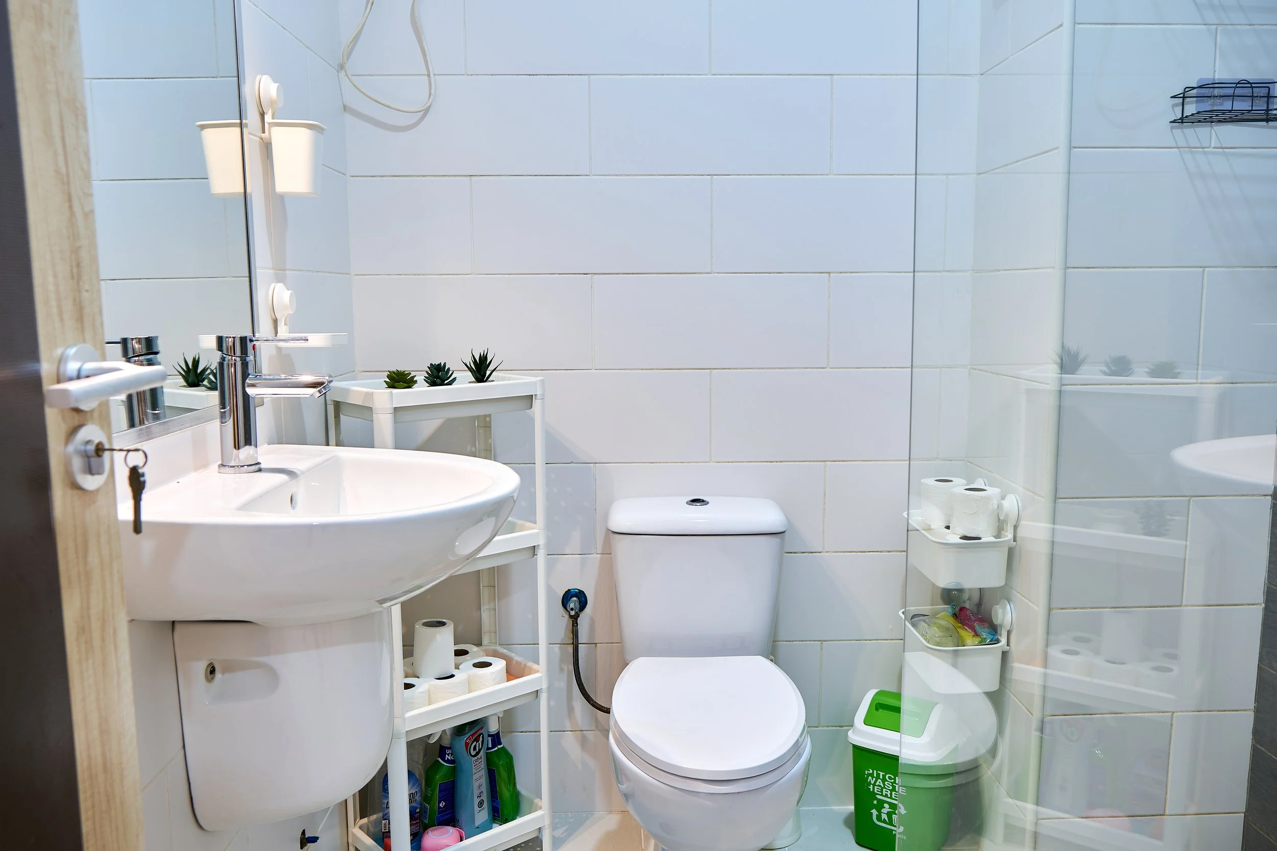 A small bathroom with white tiled walls, showing a white sink with a modern chrome faucet, a white toilet, and a small white shelf with plants. There is a mirror above the sink and a black wire basket on the wall. Toilet paper rolls are organized on 