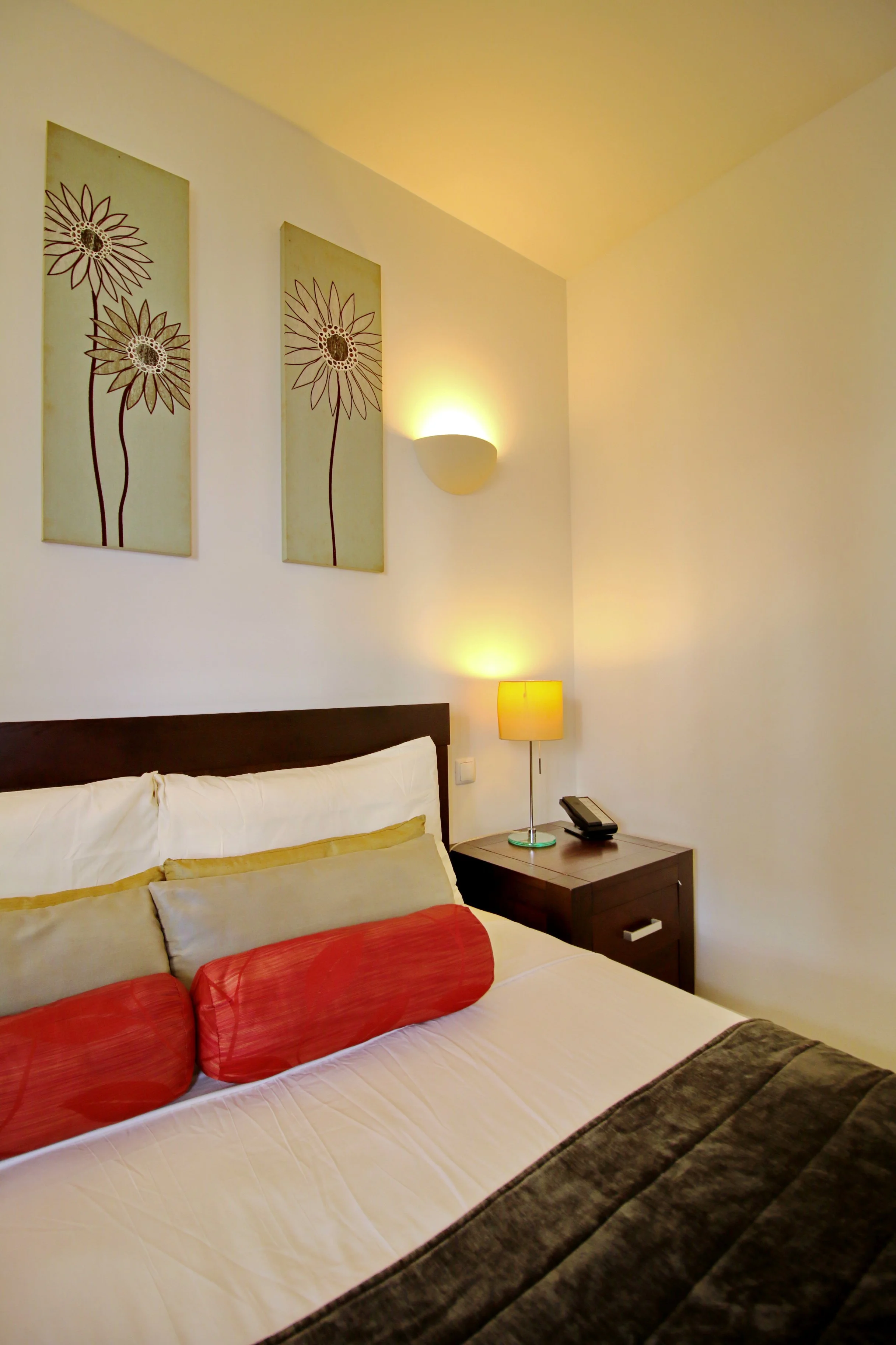 A neatly made bed with white sheets, beige pillows, and red decorative pillows in a hotel room. The headboard is dark wood. To the right, there is a nightstand with a lamp, a telephone, and a notepad. Wall art depicting flowers and a wall sconce ligh